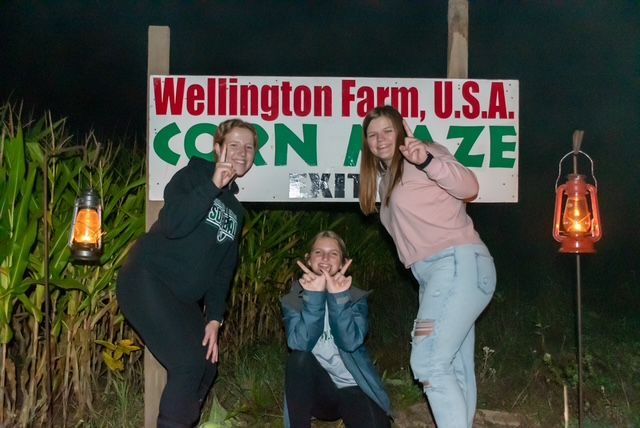 Three girls are posing in front of a sign that says wellington farm u.s.a. corn maze