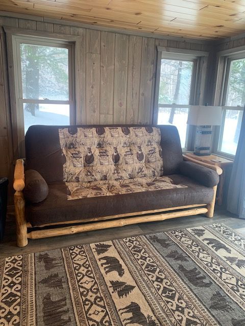 A brown futon with a patterned cover sits in a rustic cabin, with windows and a patterned rug.