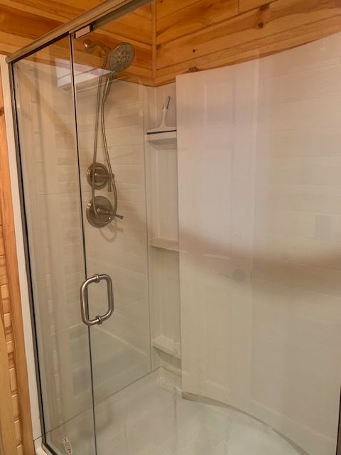 Shower stall with glass door, fixtures, and white curved wall, set in wood-paneled room.