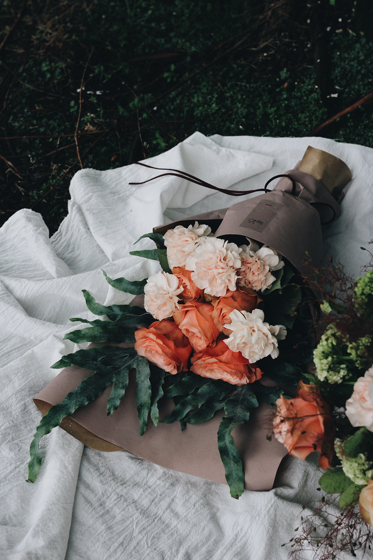 Bouquet of peach and cream flowers wrapped in brown paper, resting on a white cloth on grass.