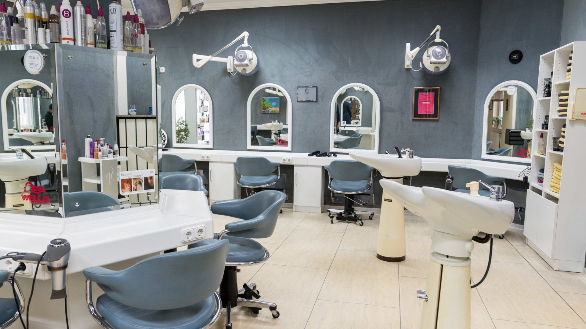 Inneneinrichtung eines Friseursalons: blaue Stühle, weiße Stationen, gewölbte Spiegel, Geräte und Produkte.