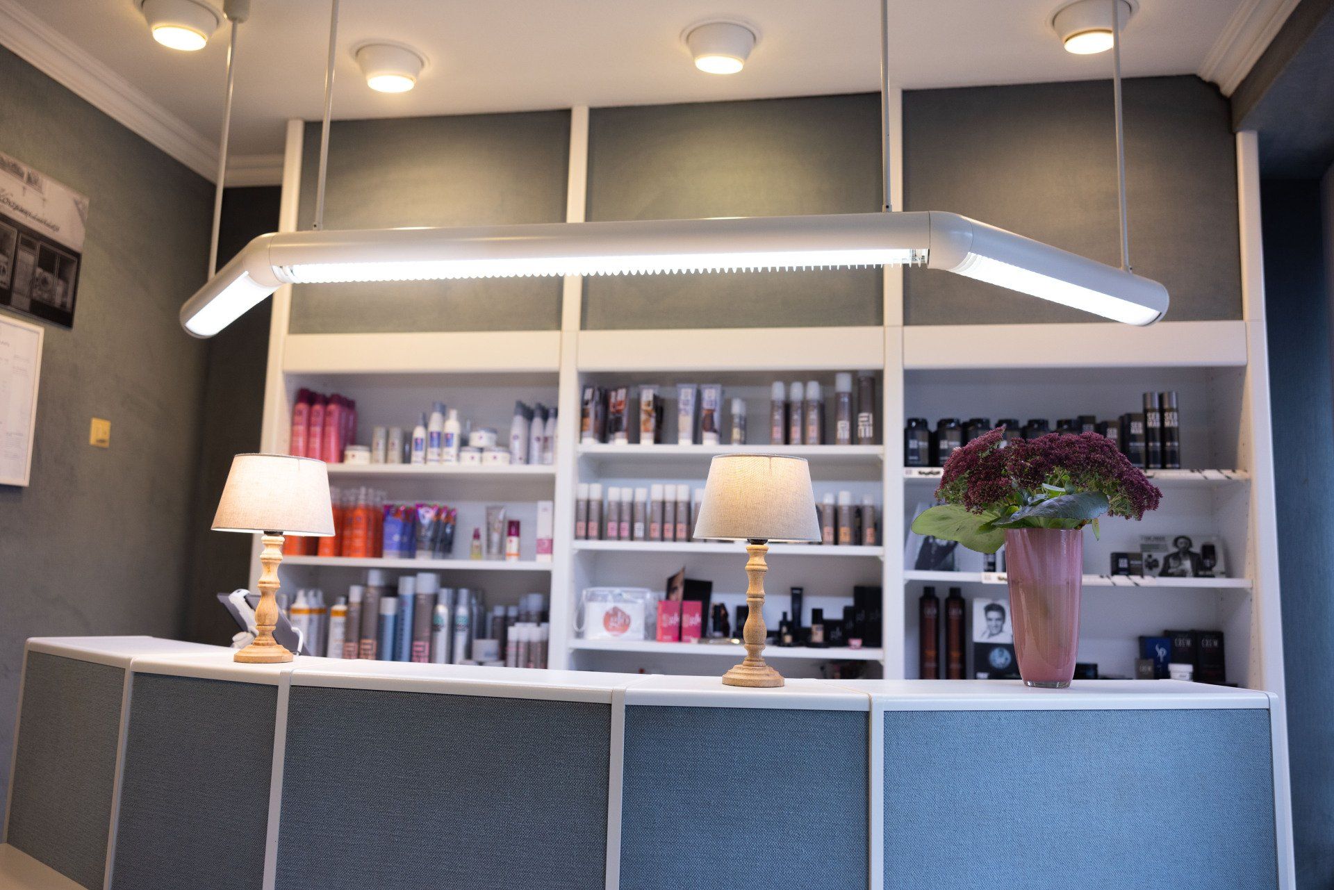 Empfangsbereich eines Friseursalons mit weißen Regalen, Produkten, Lampen und dekorativer Vase.