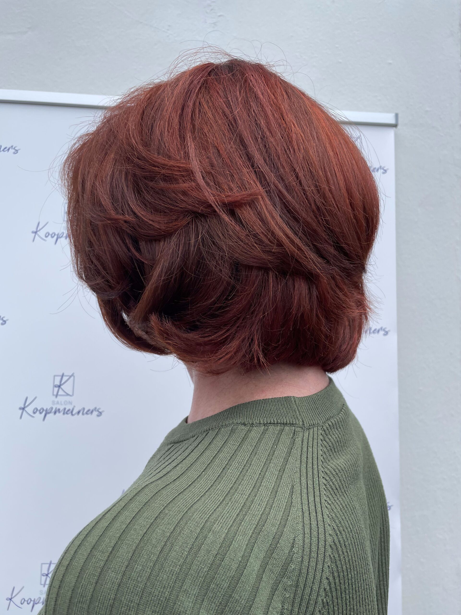 Frau mit roten Haaren und grünem Pullover, Seitenansicht, Haare in Stufen gestylt.