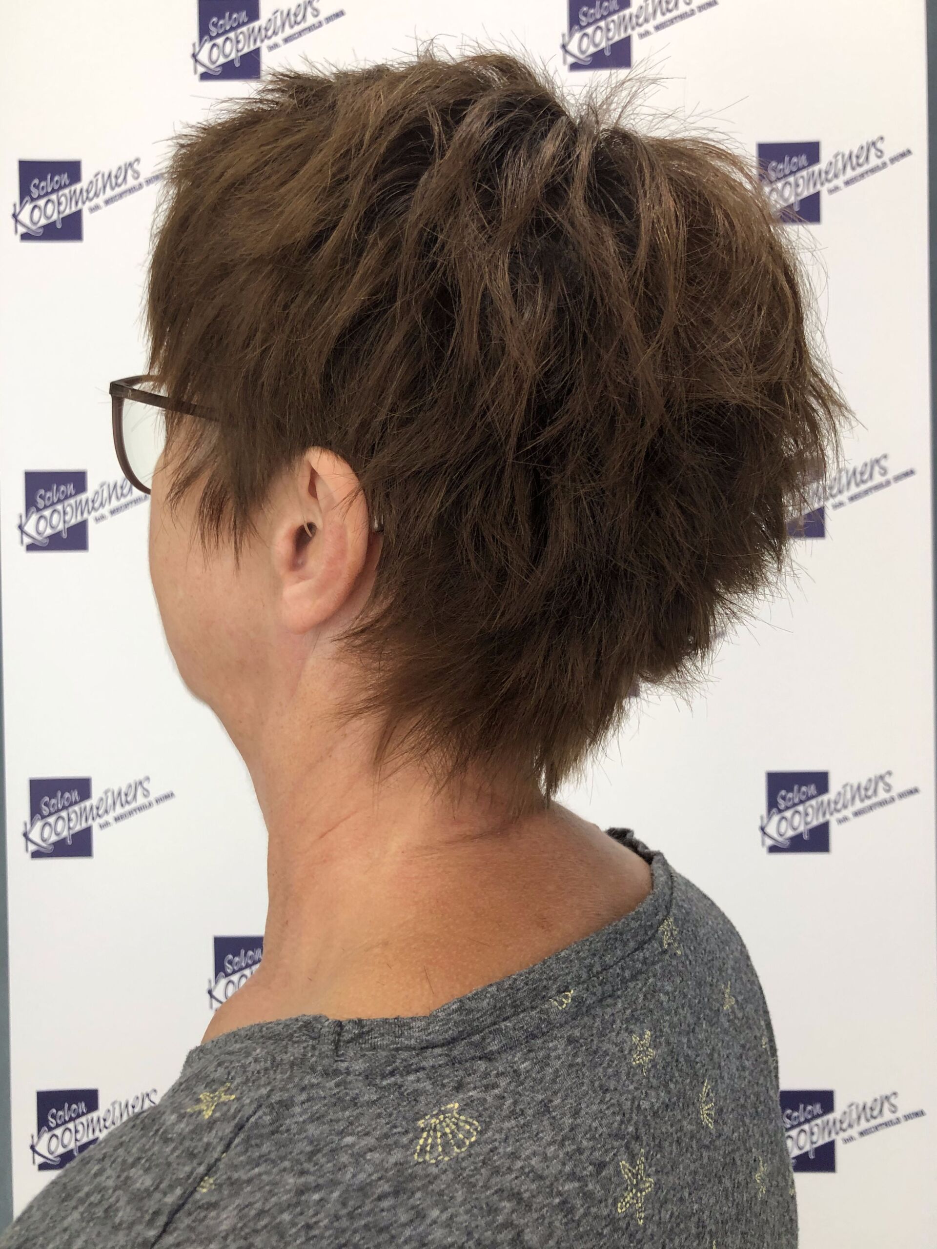 Frau mit braunem, strukturiertem Kurzhaar, von hinten; trägt eine Brille und ein graues Hemd.