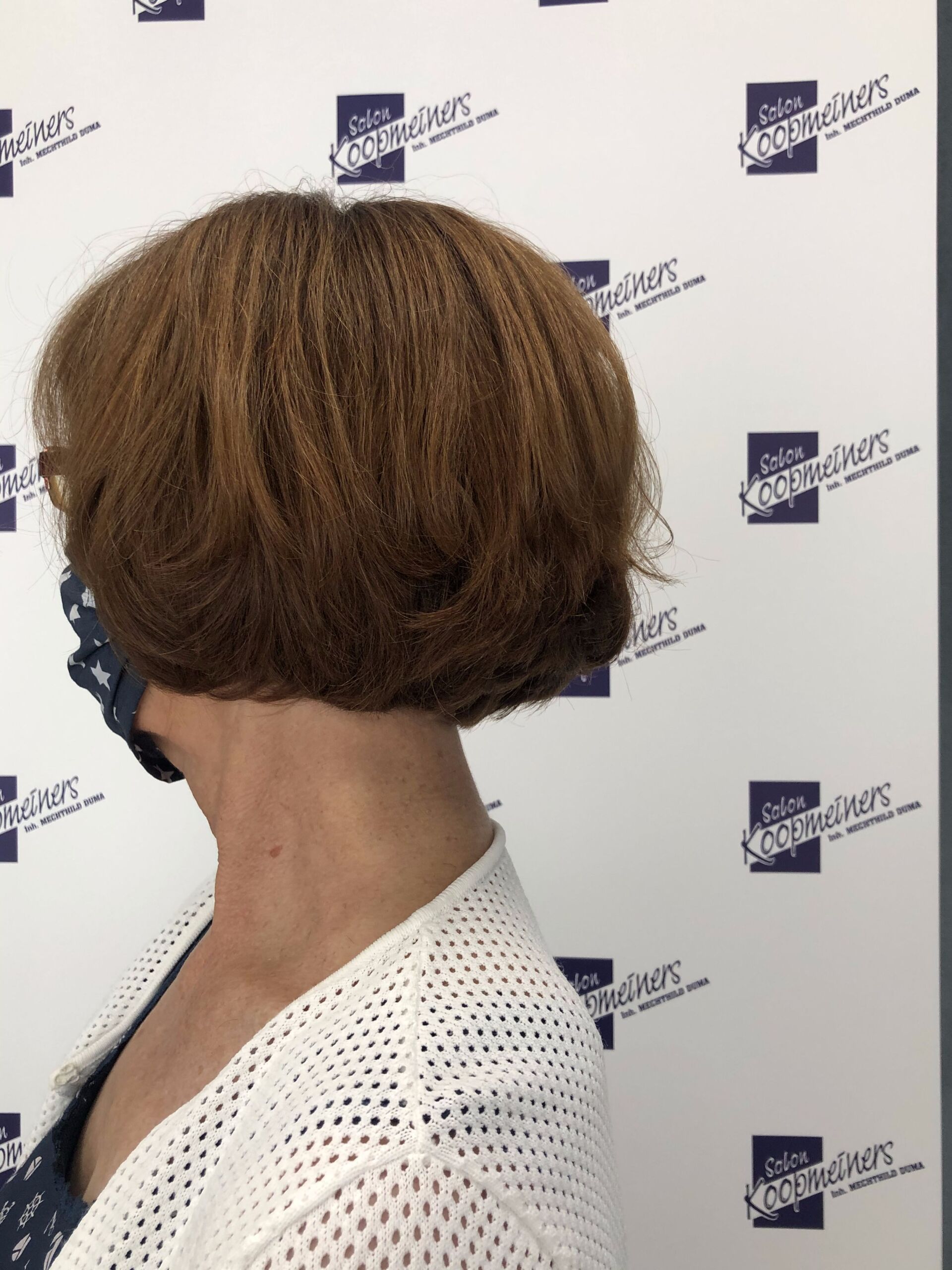 Frau mit kastanienbraunem Bob-Haarschnitt, Maske und weißer Strickjacke vor gemustertem Hintergrund.