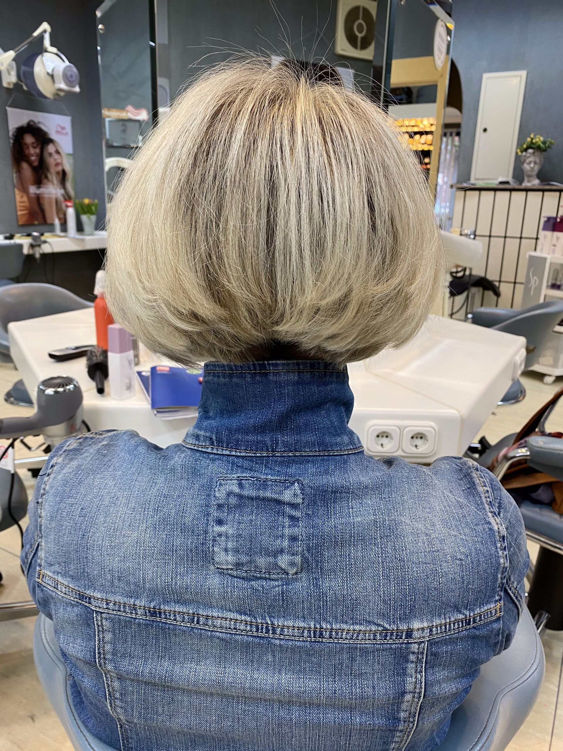 Frau mit blonder Bobfrisur und blauer Jeansjacke in einem Salon.