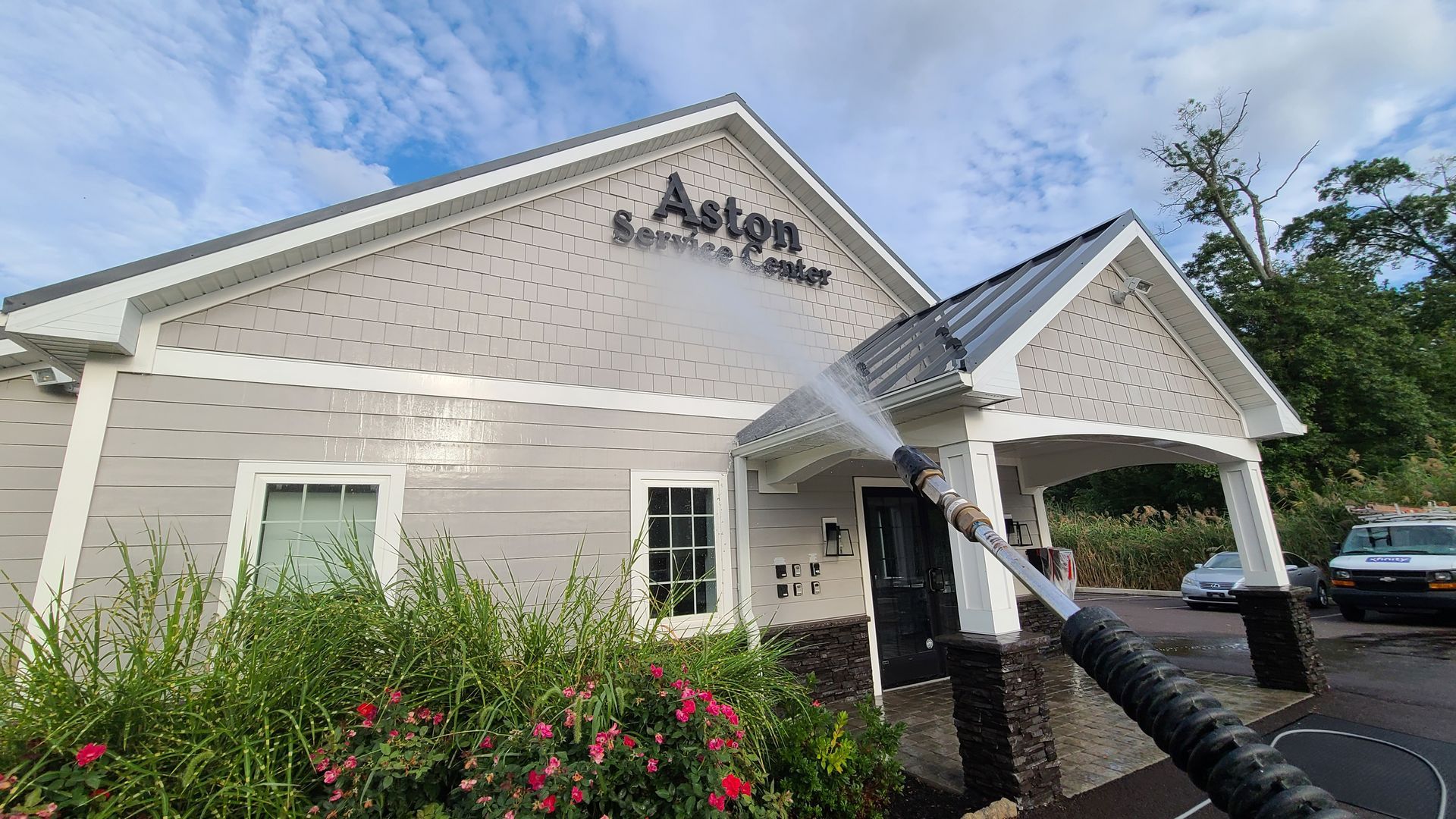 Exterior of a beige building with black awning, white trim, and flowering bushes under a blue sky
