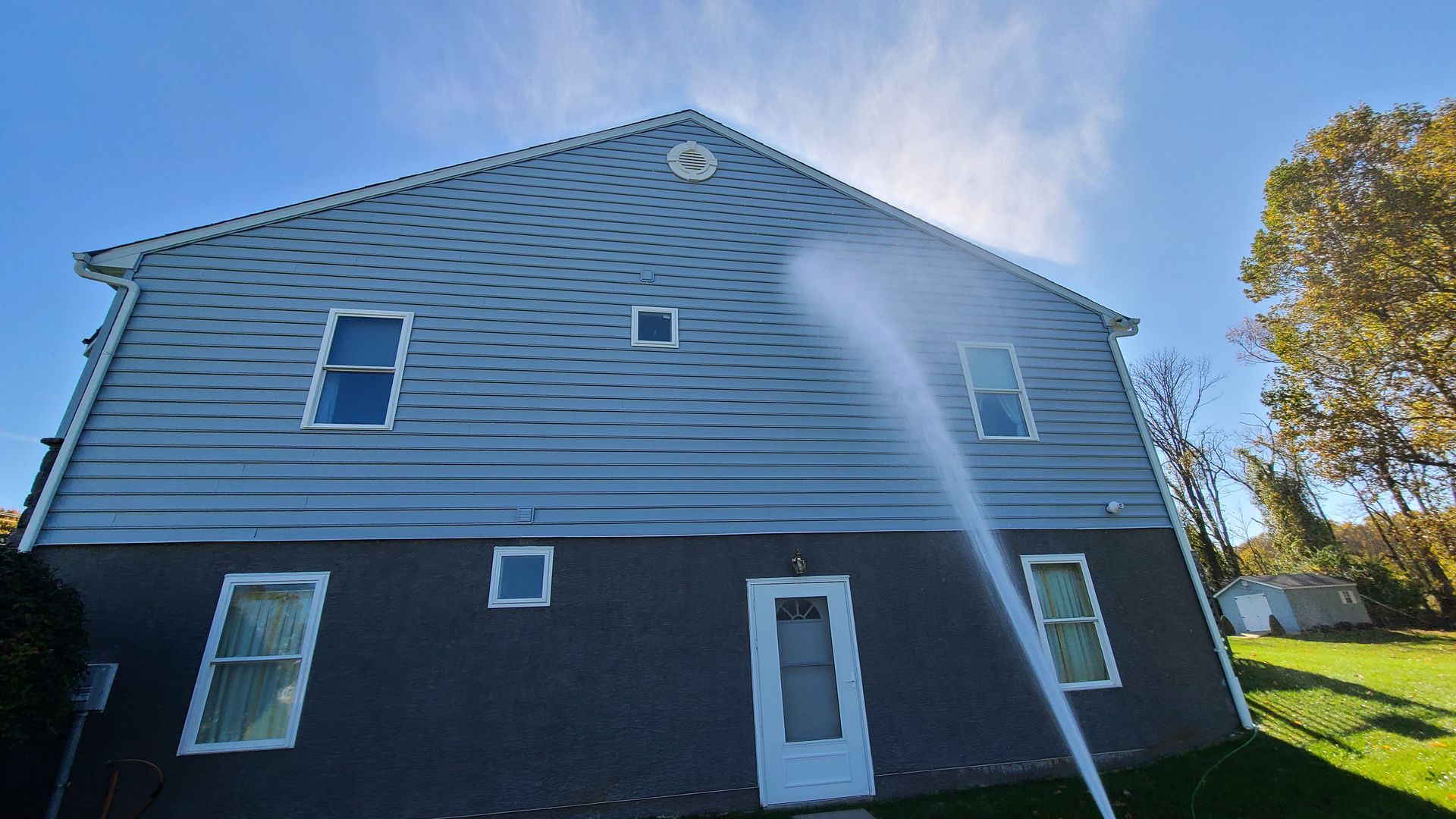 Pressure washing a two-story blue-gray house with white trim on a sunny day.