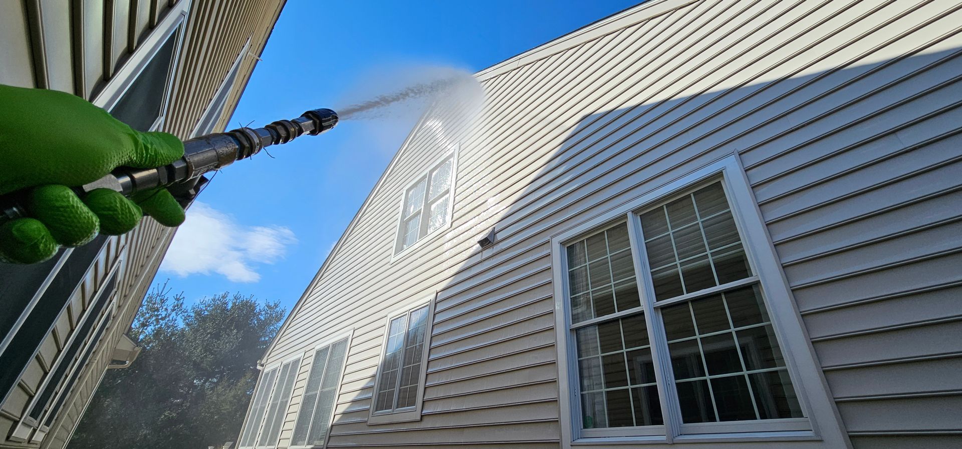 Green-handled pressure washer spraying water on white house siding and windows outdoors