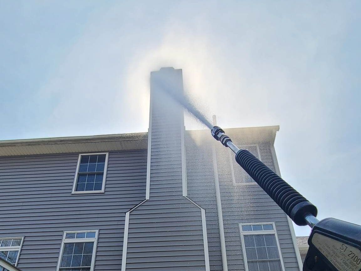 Pressure-washing the side of a house, with strong spray hitting the upper siding near the roofline.