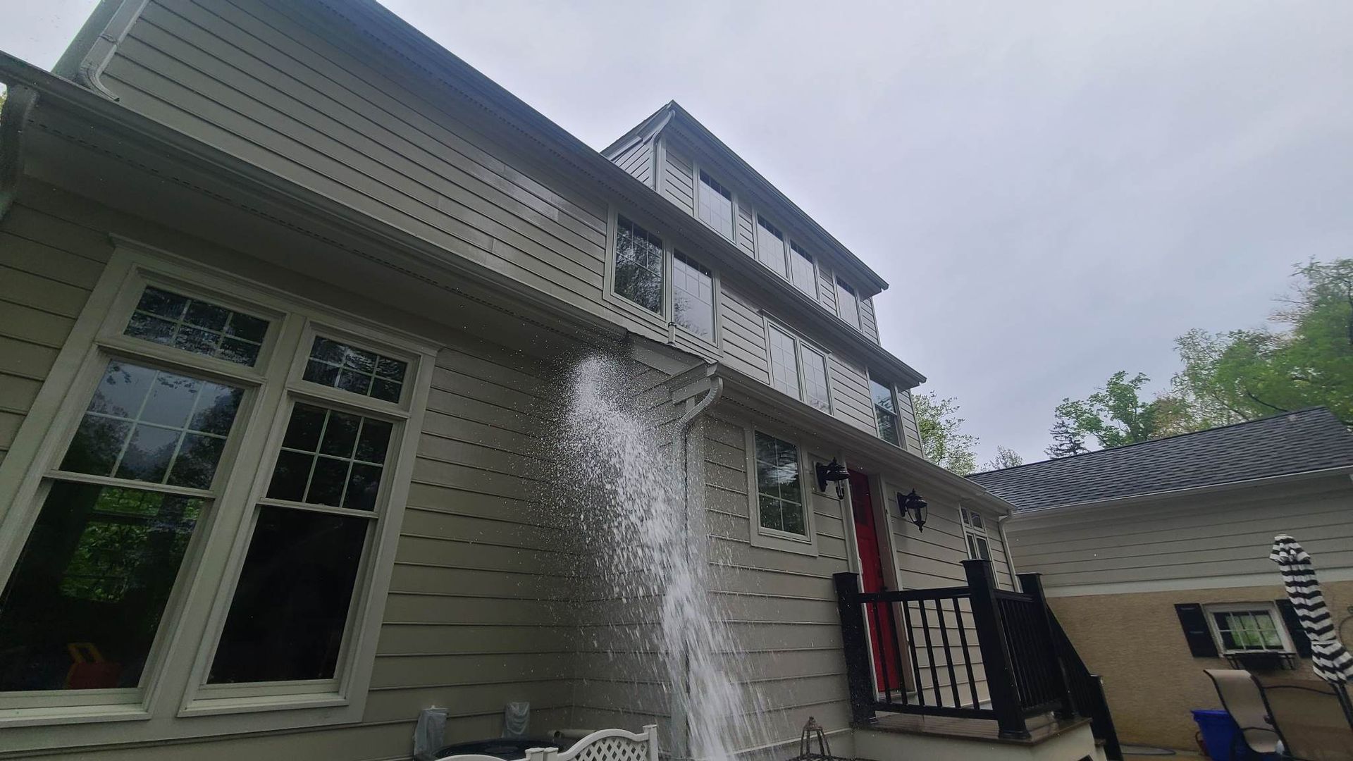 Water sprays from a building's gutter during cleaning. Light-colored siding, windows, and red door. Cloudy sky.