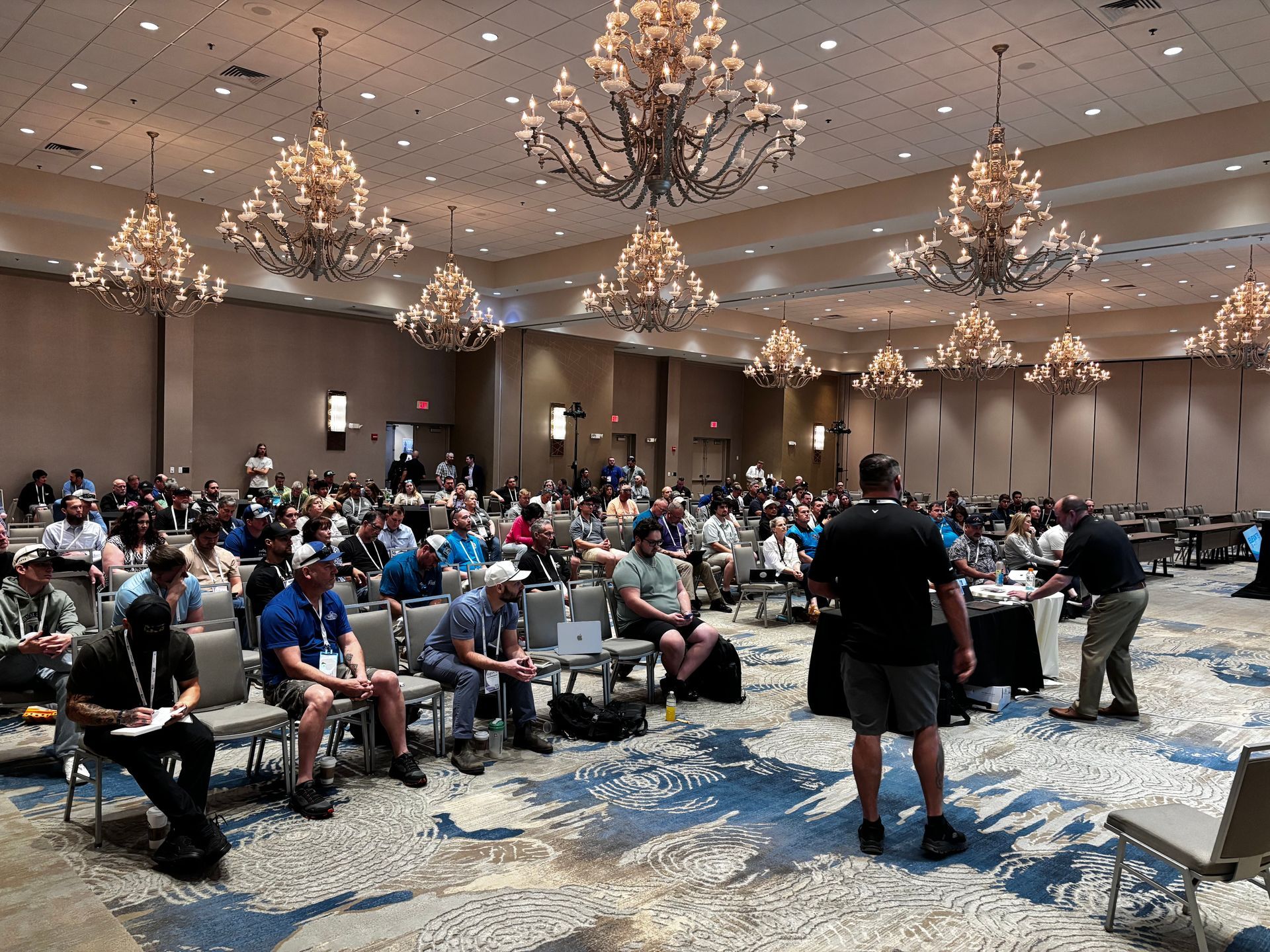 Large conference room with seated audience and a speaker at the front under chandeliers.