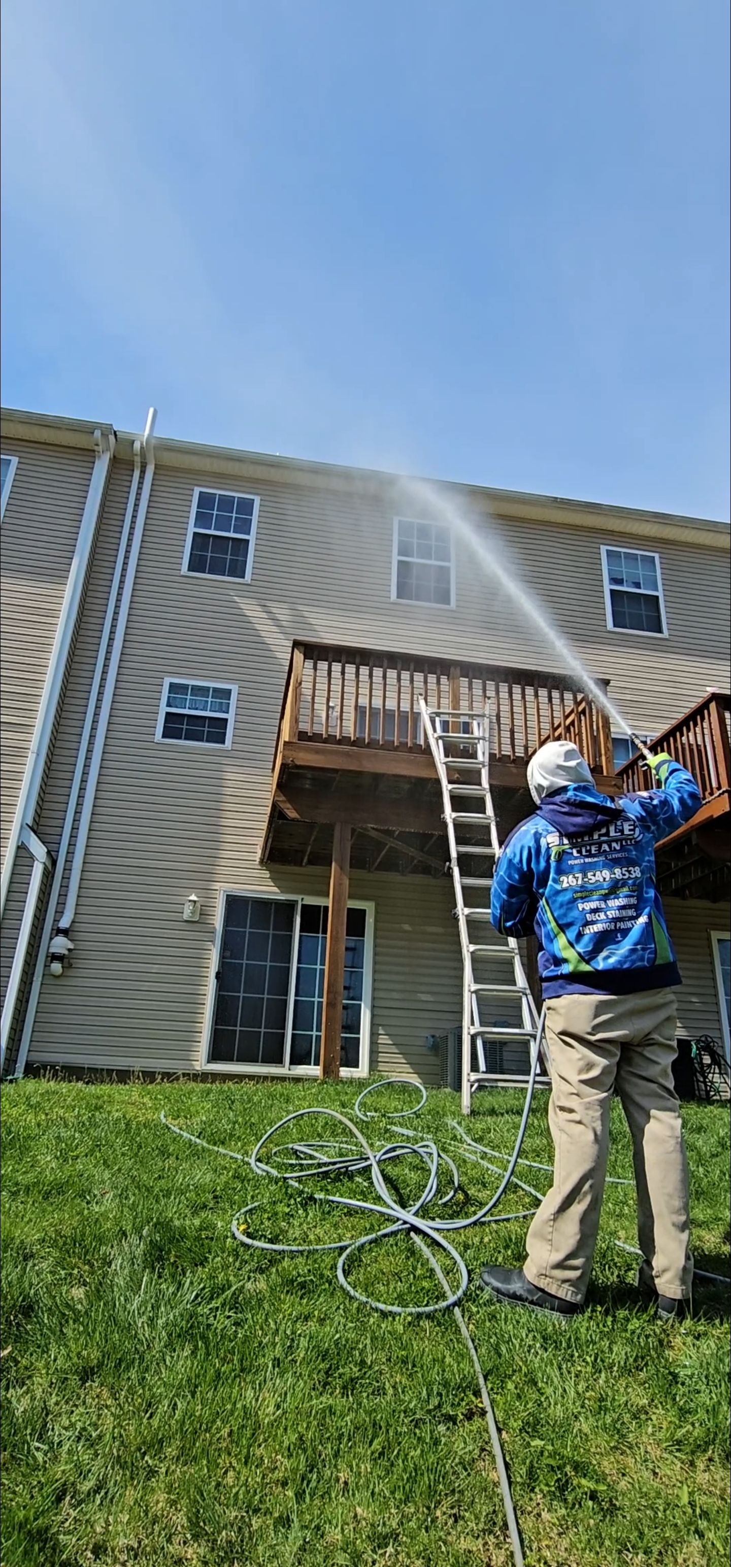 Person in blue shirt power washing a house; outdoor daytime.