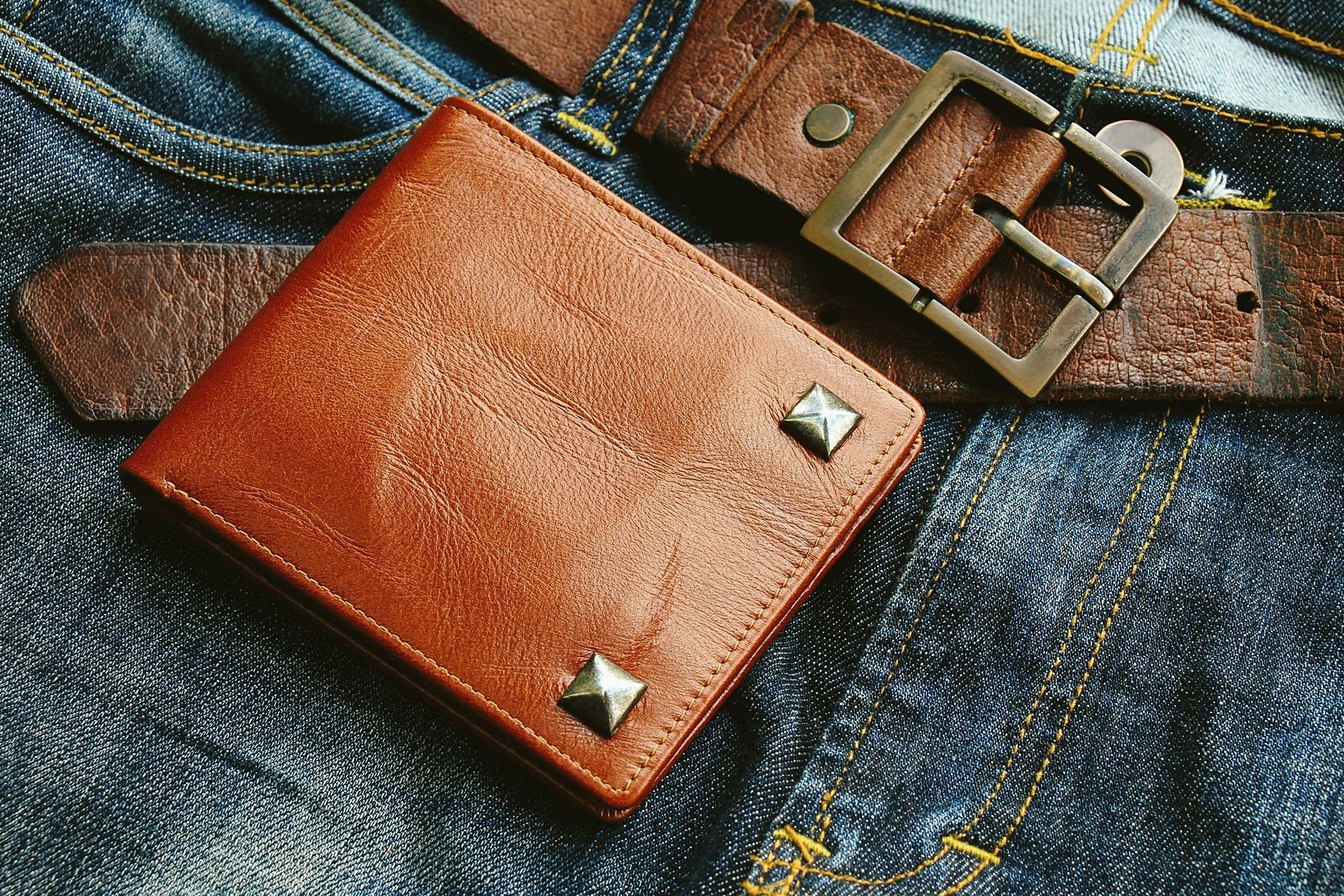 Brown leather wallet with studded corners, resting on a brown belt over denim jeans.