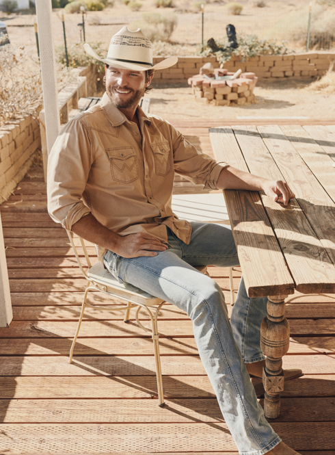 Man in plaid shirt, cowboy hat, and bandana, leaning against a wooden wall with hands in pockets.