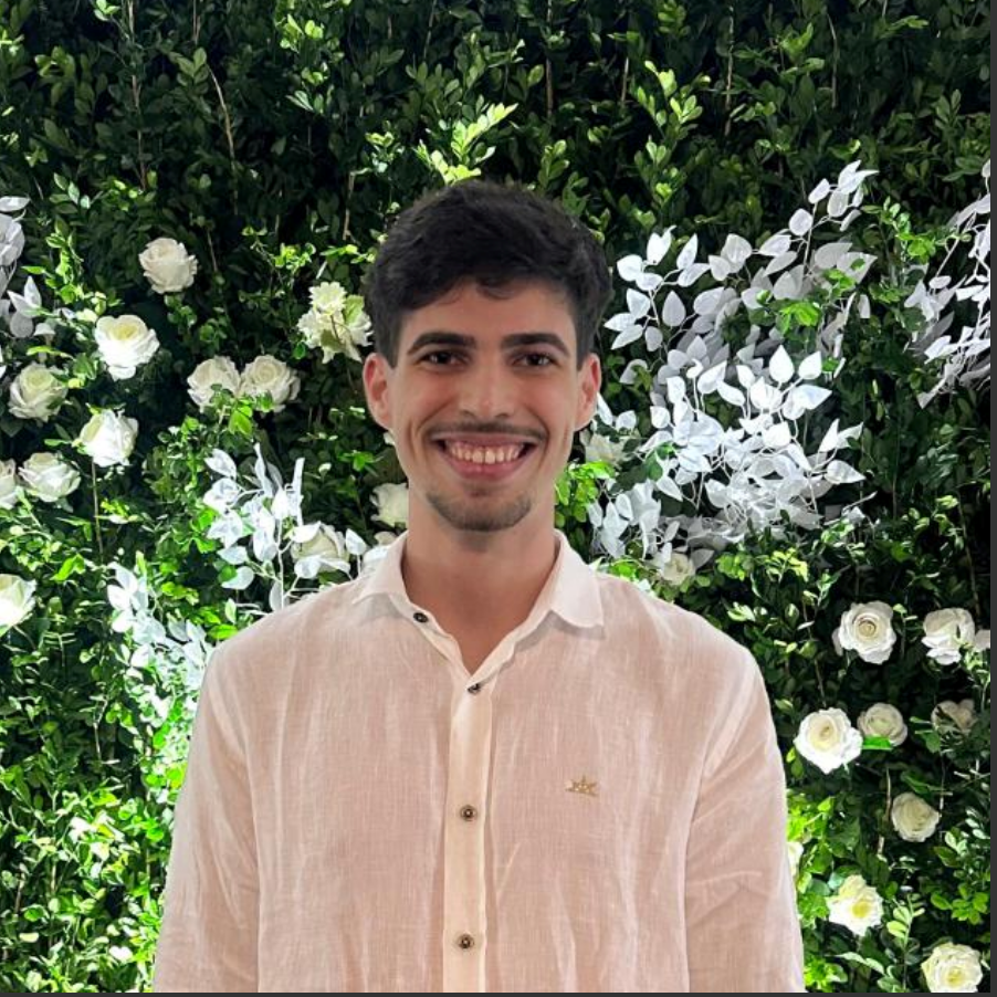 Homem sorridente com camisa rosa claro em pé diante de um exuberante cenário floral verde e branco.