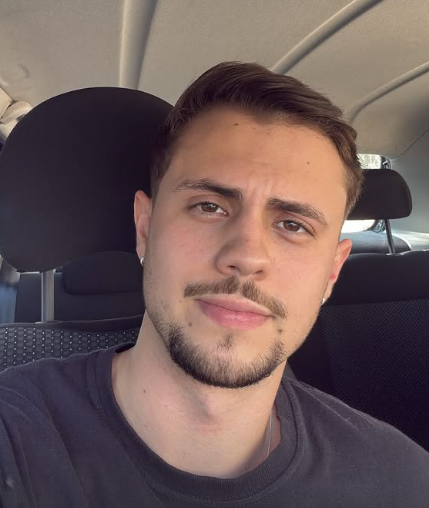 Homem sentado em um carro, olhando para a câmera com uma expressão neutra.