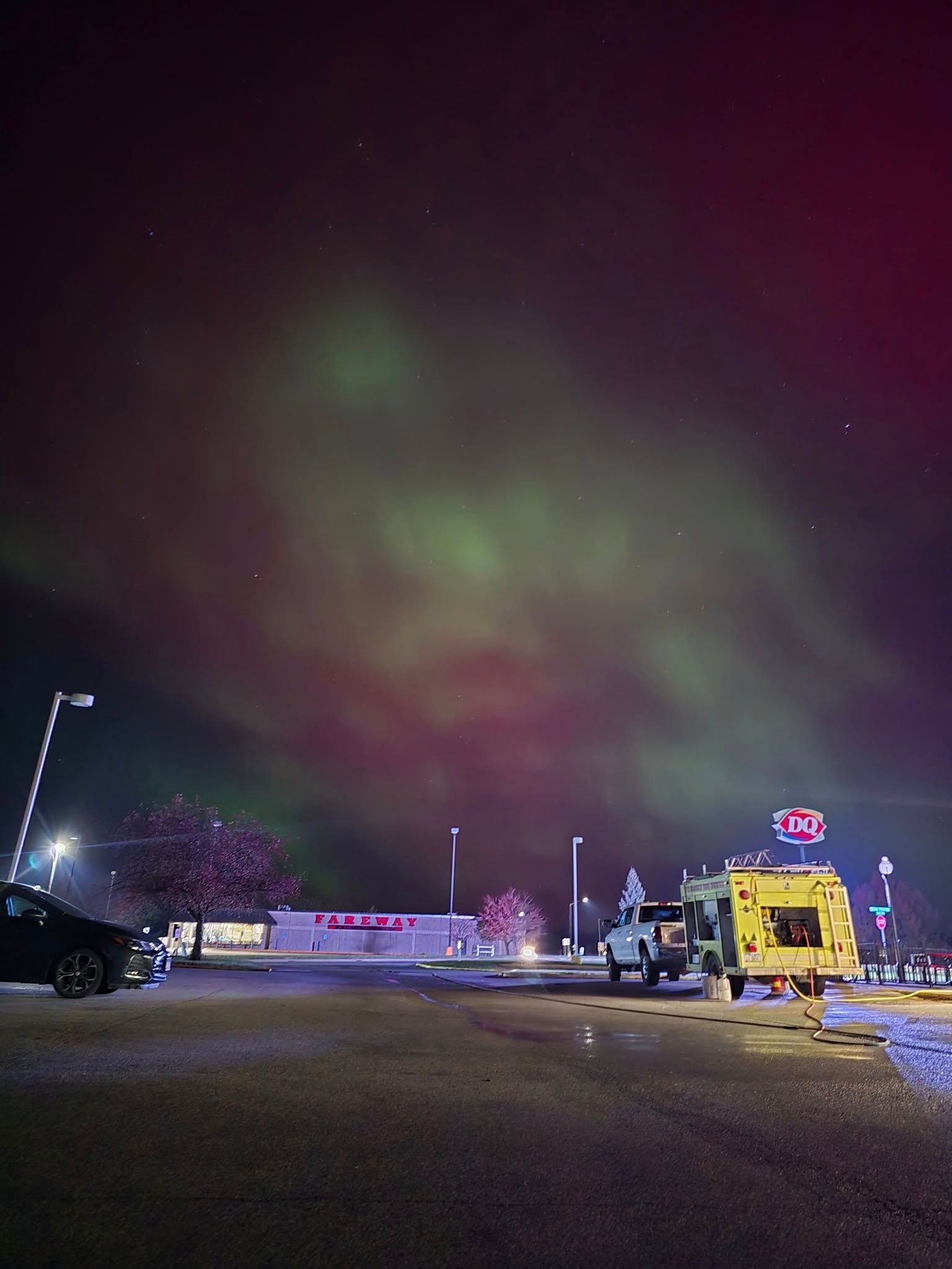 Green and red aurora borealis lights fill the night sky above a parking lot with parked vehicles and a building.