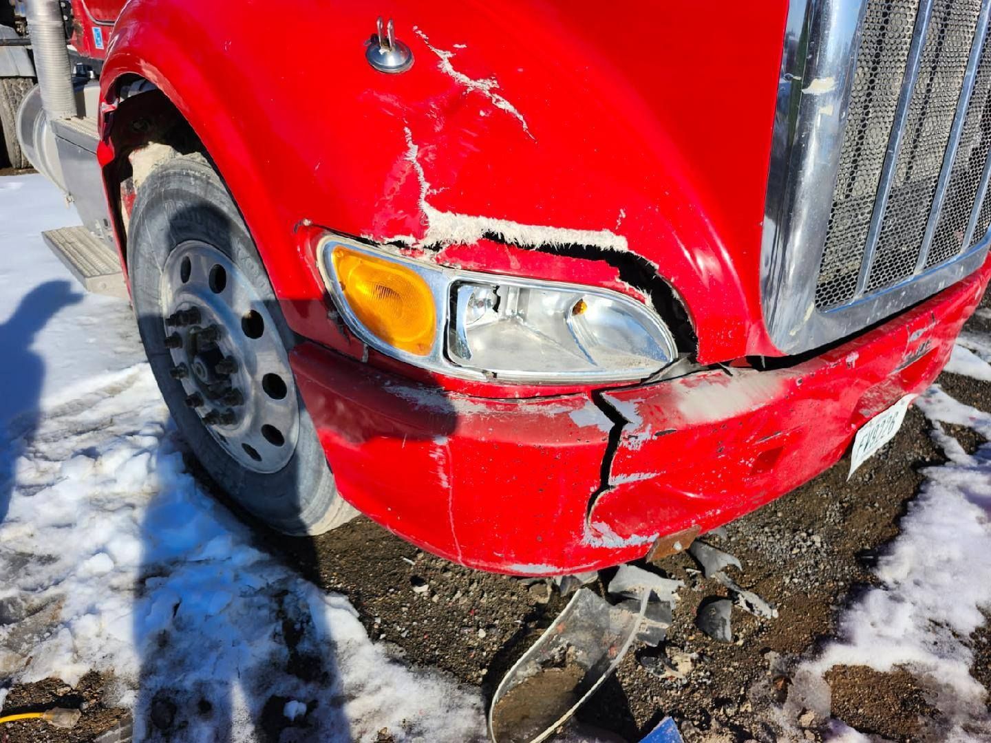 A damaged red truck hood and bumper show a crack, broken light assembly, and paint peeling, set against snowy ground.