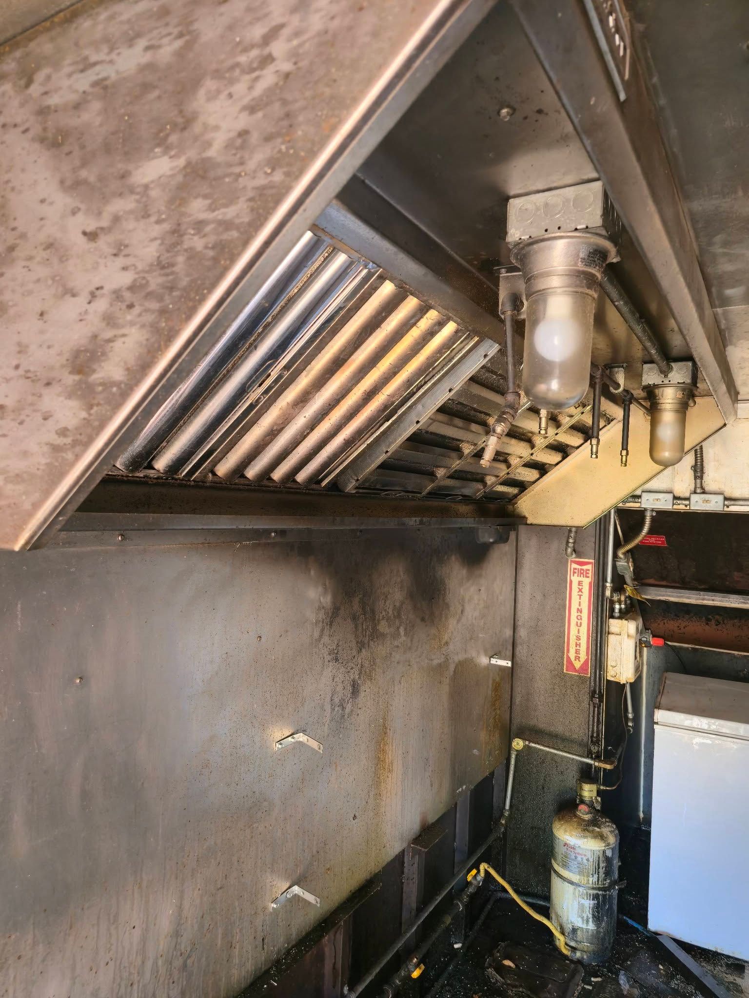 A dirty stainless steel commercial kitchen exhaust hood with grease-stained walls and a light fixture.
