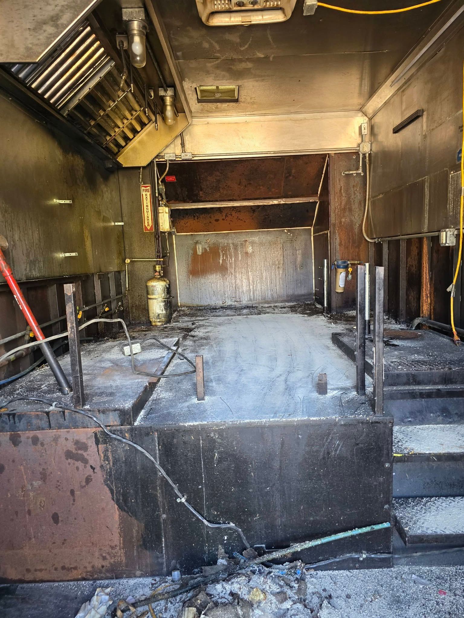 The charred, empty interior of a food truck after a fire, showing blackened walls, equipment frames, and debris.