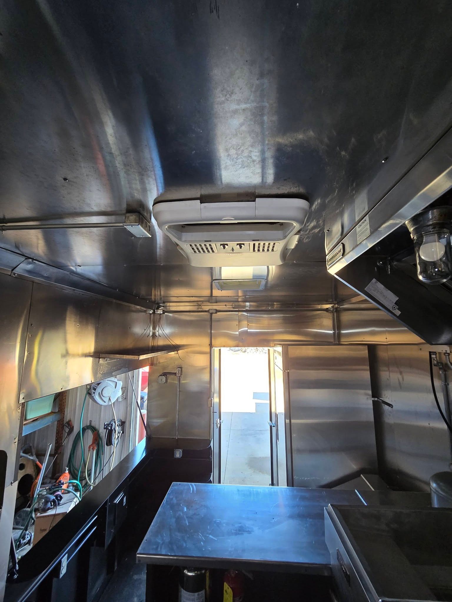 Interior view of a food truck with metallic walls, a ceiling-mounted ventilation unit, prep counters, and an open doorway.