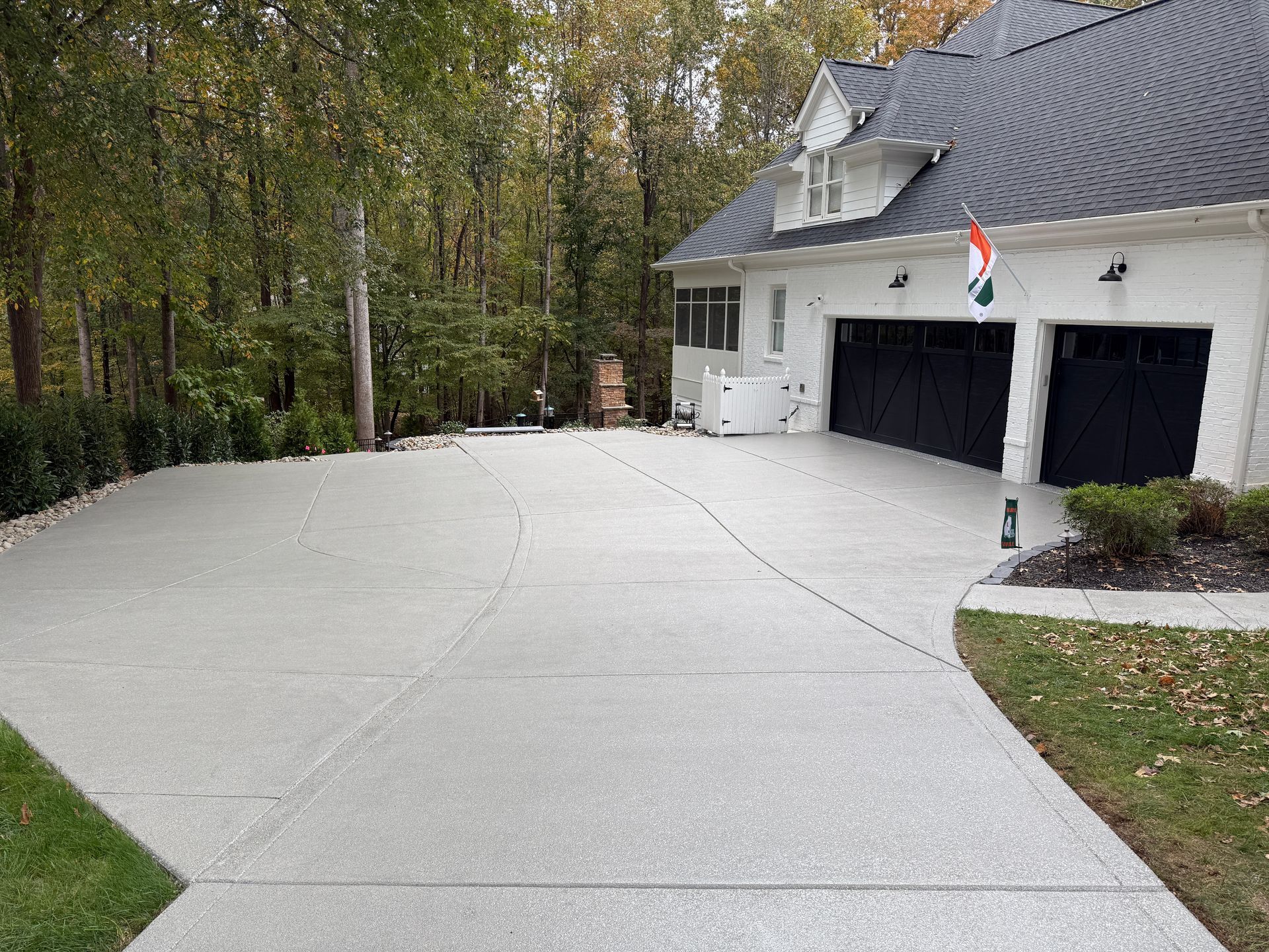 Large concrete driveway leading to a white house with two black garage doors and a small flag, surrounded by trees