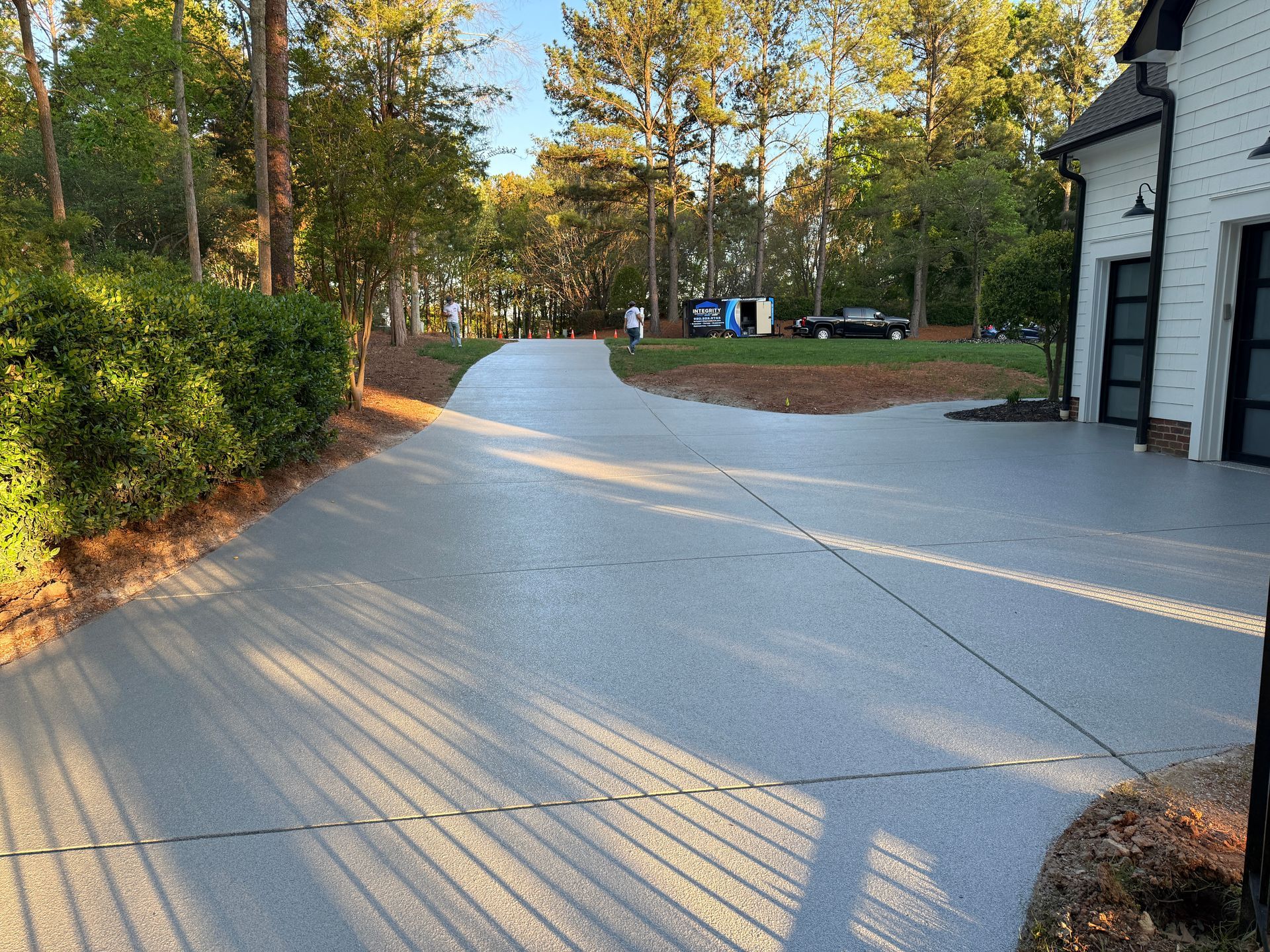 Curved concrete driveway beside a house, lined with trees and bushes in warm sunlight.