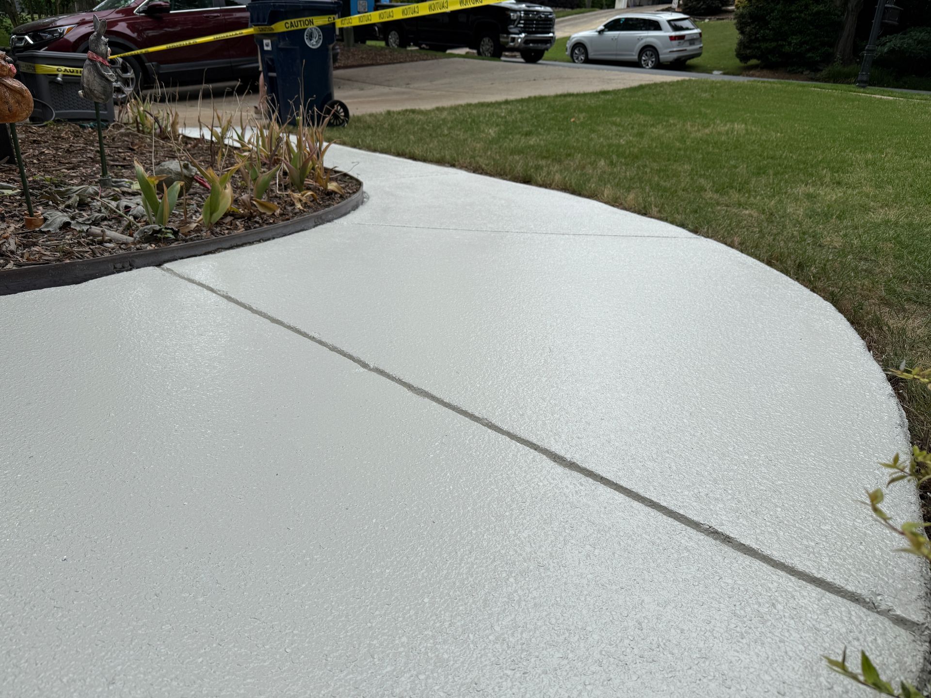 Freshly poured light gray concrete driveway next to a lawn and flower bed, with yellow caution tape in the background