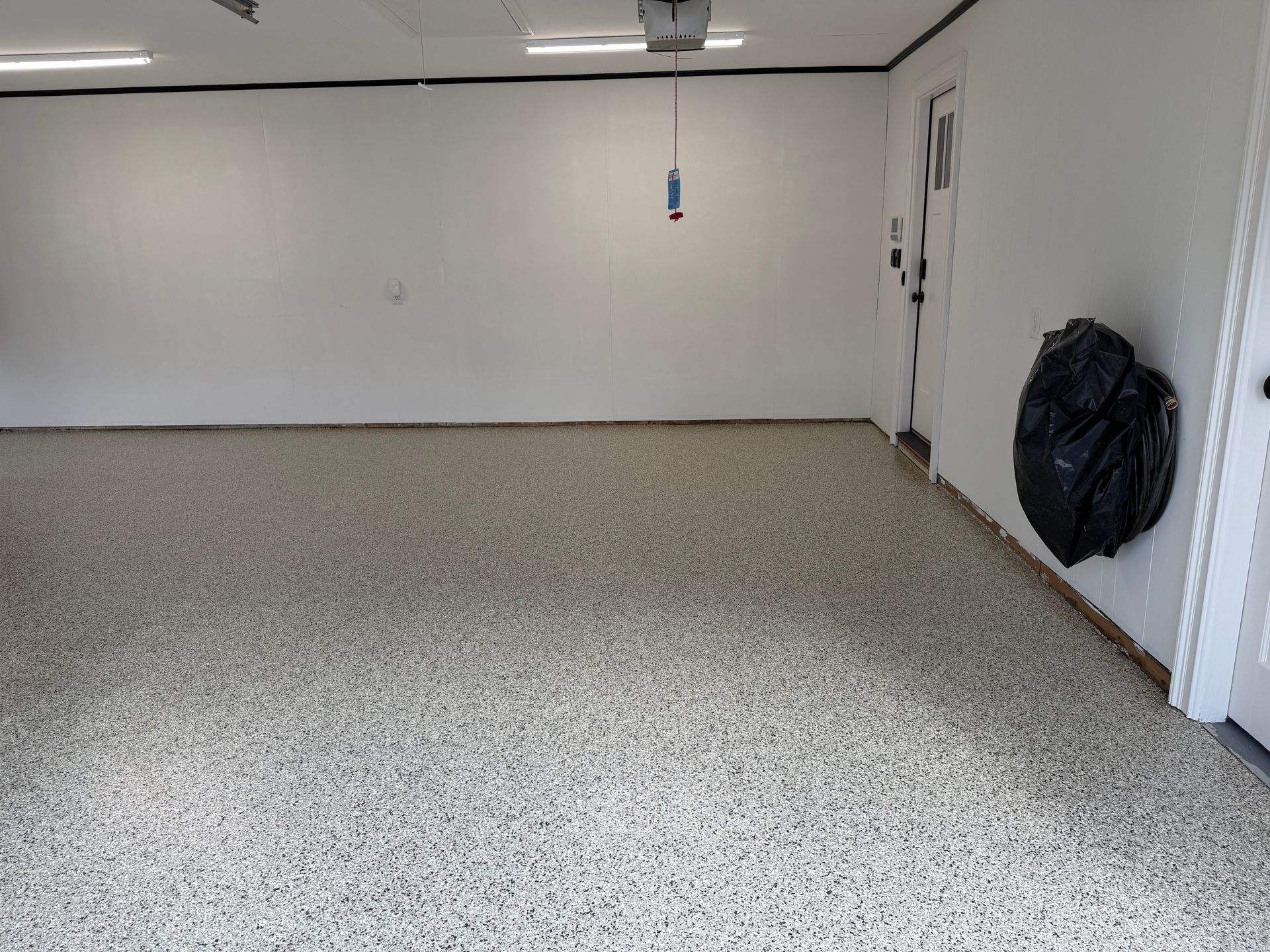 Empty garage with white walls, speckled floor, and a black trash bag by the right wall.