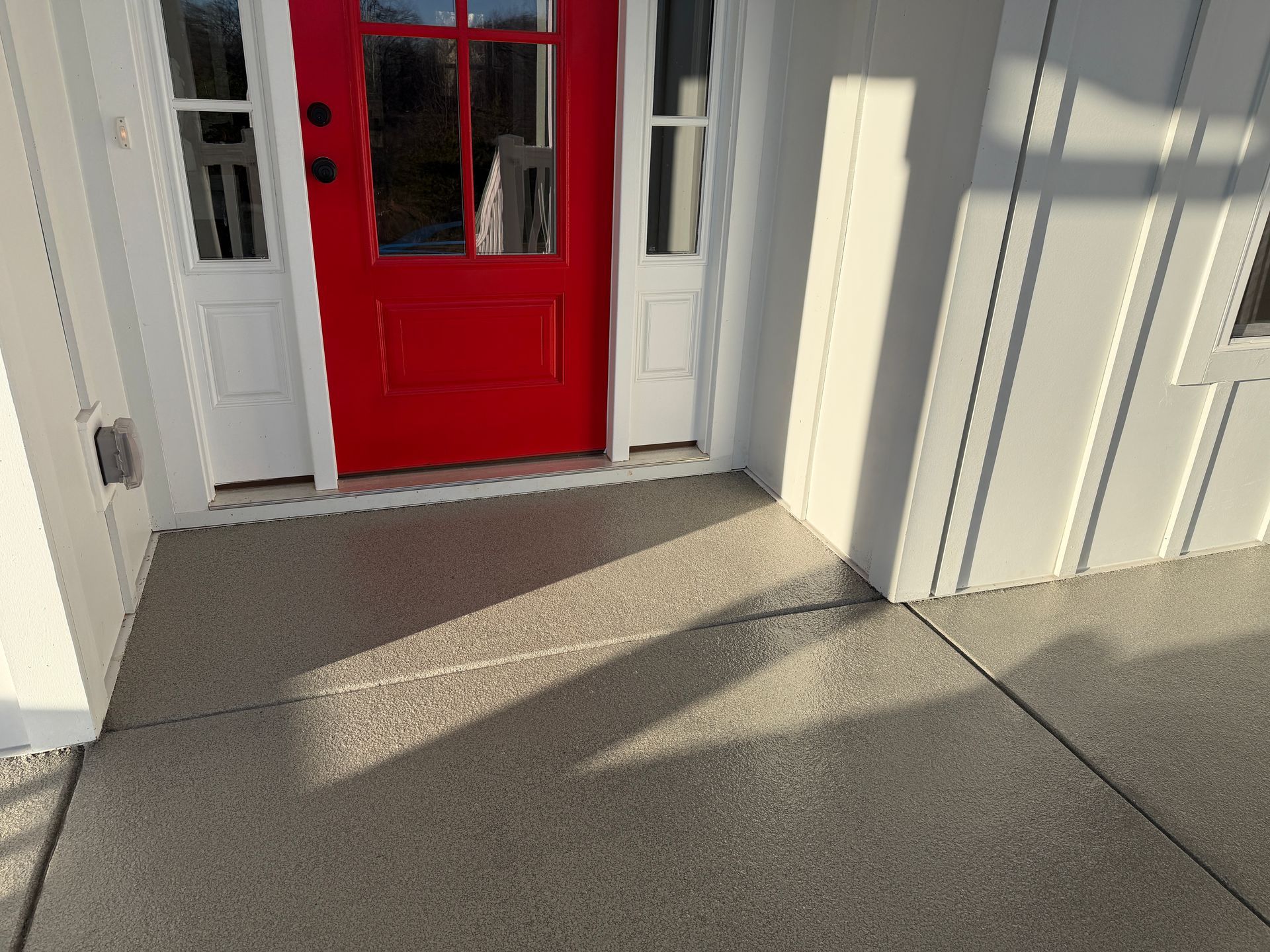 Front porch with a bright red door, white siding, and sunlight casting long shadows on the floor.