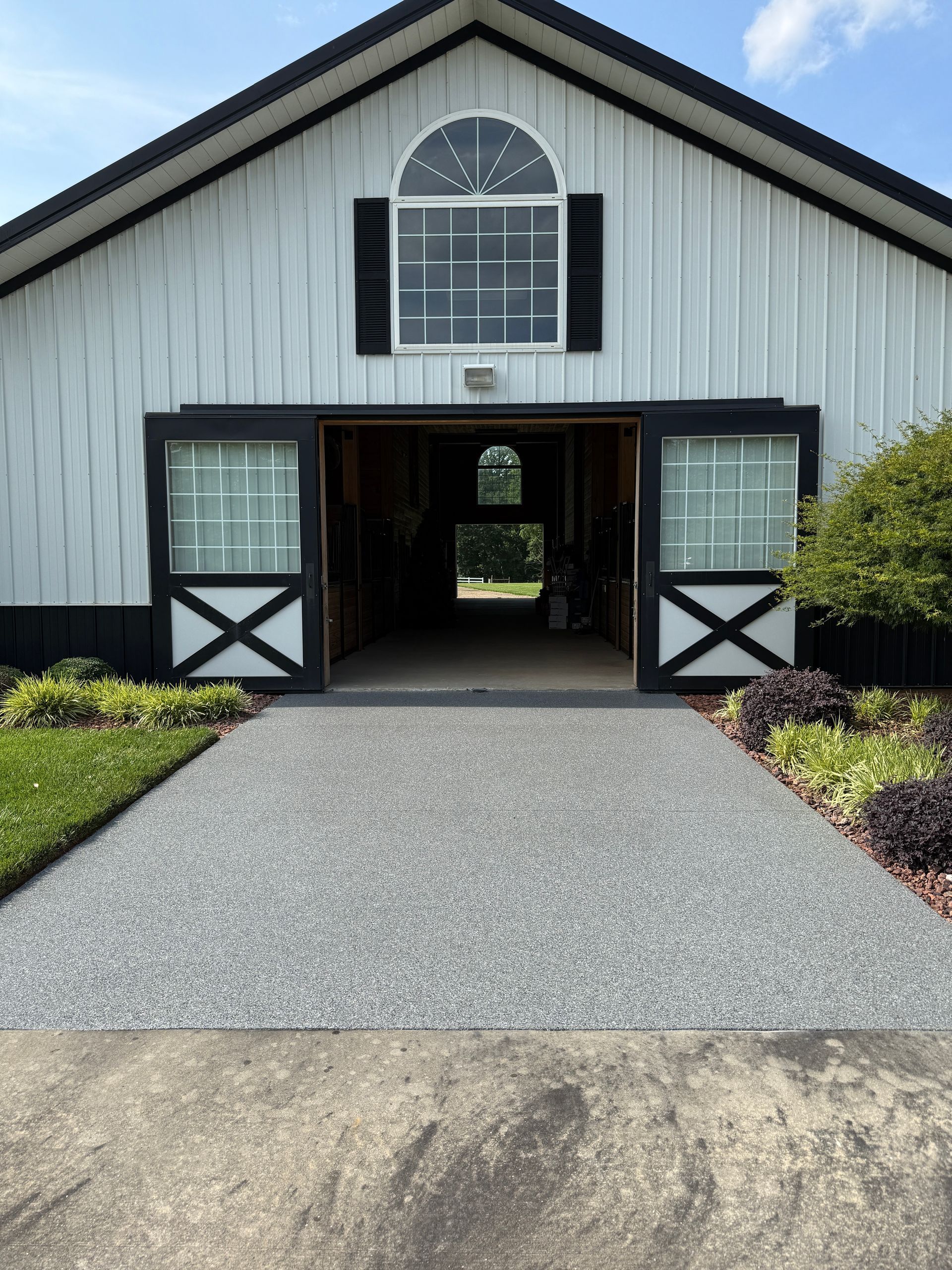 White barn with black trim and open doors at the end of a gravel driveway, framed by flower beds and shrubs