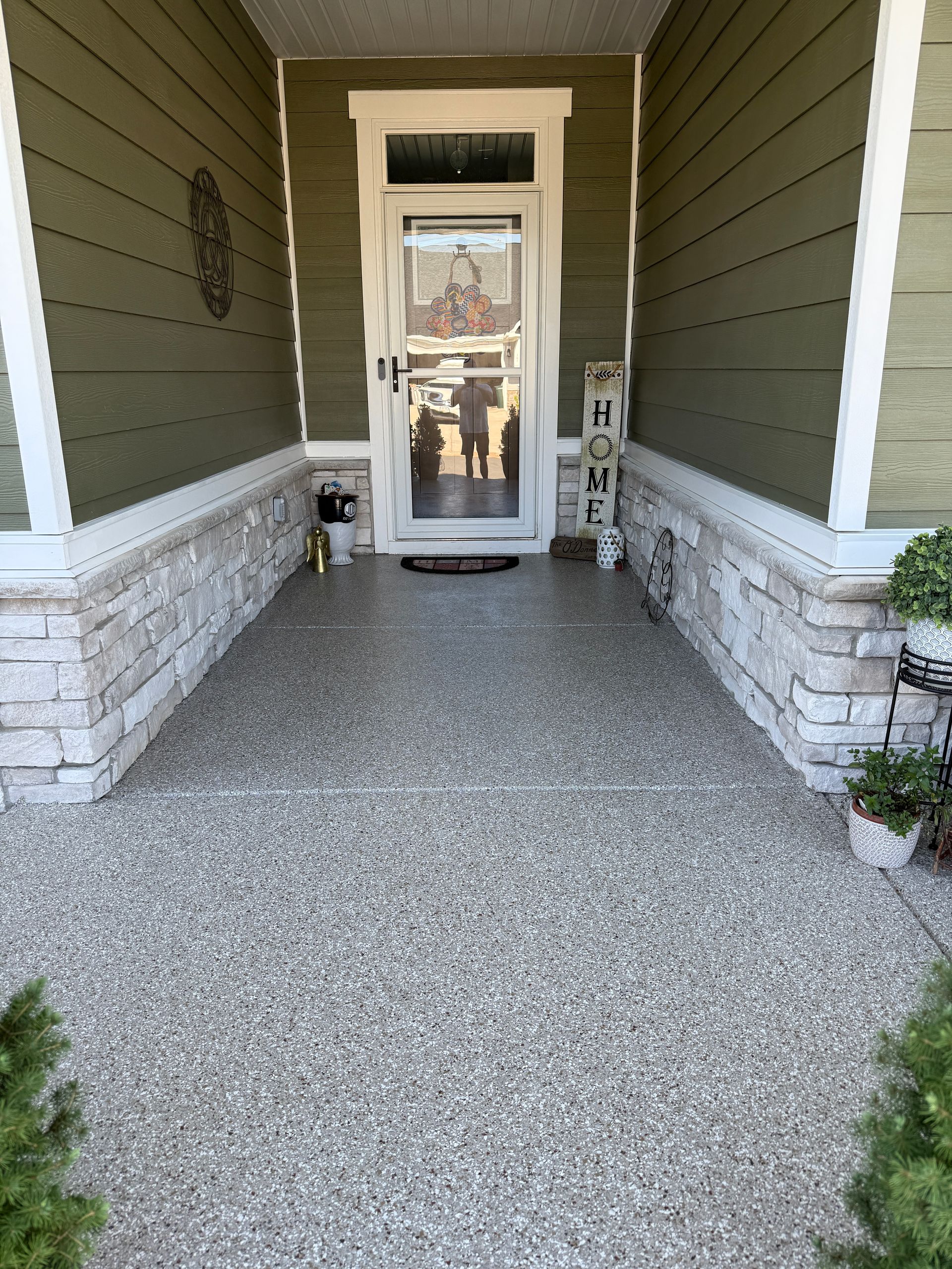 Covered front porch with speckled concrete floor, stone half-walls, and a glass storm door.