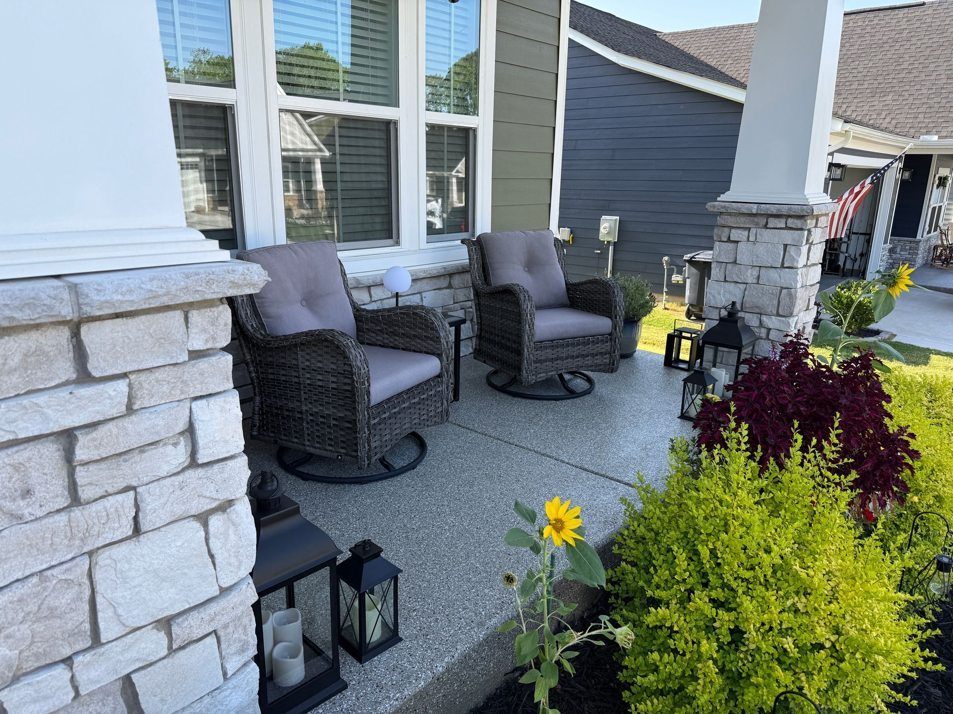 Front porch with wicker chairs, lanterns, and potted flowers beside a stone column and gray siding.