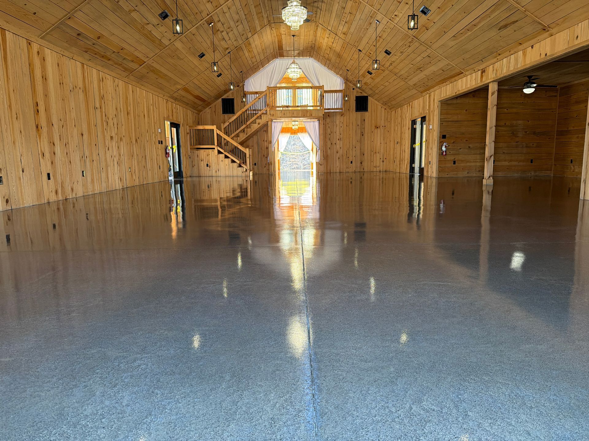Empty wooden hall with a glossy gray floor, stairs at the back, and sunlight through the central doorway.