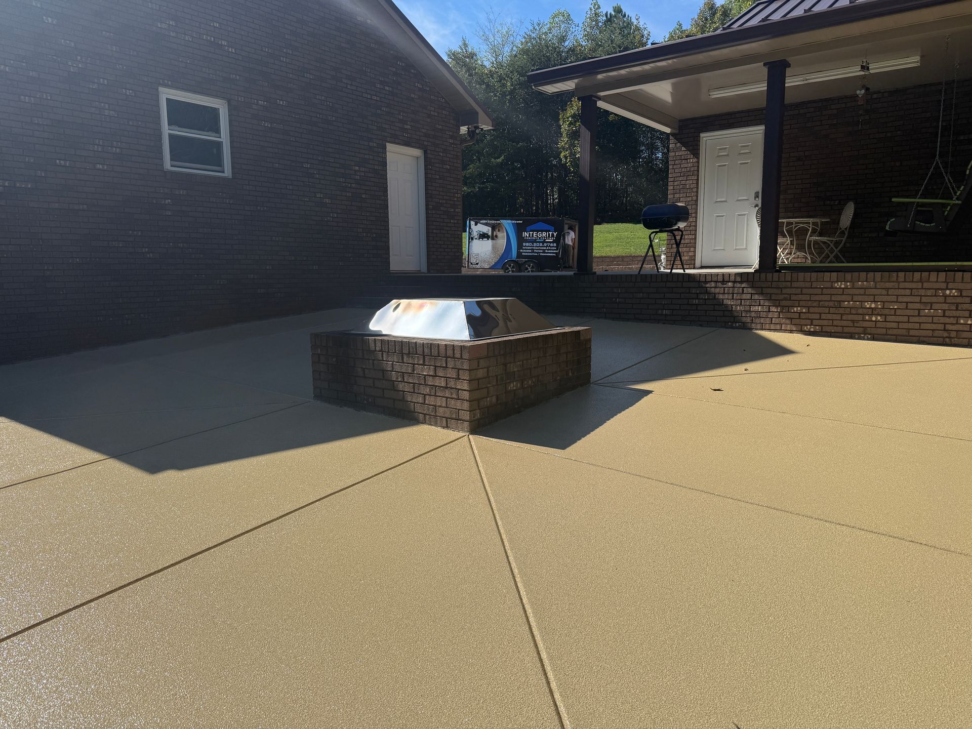 Brick fire pit on a sunlit concrete patio beside a house and covered outdoor area.