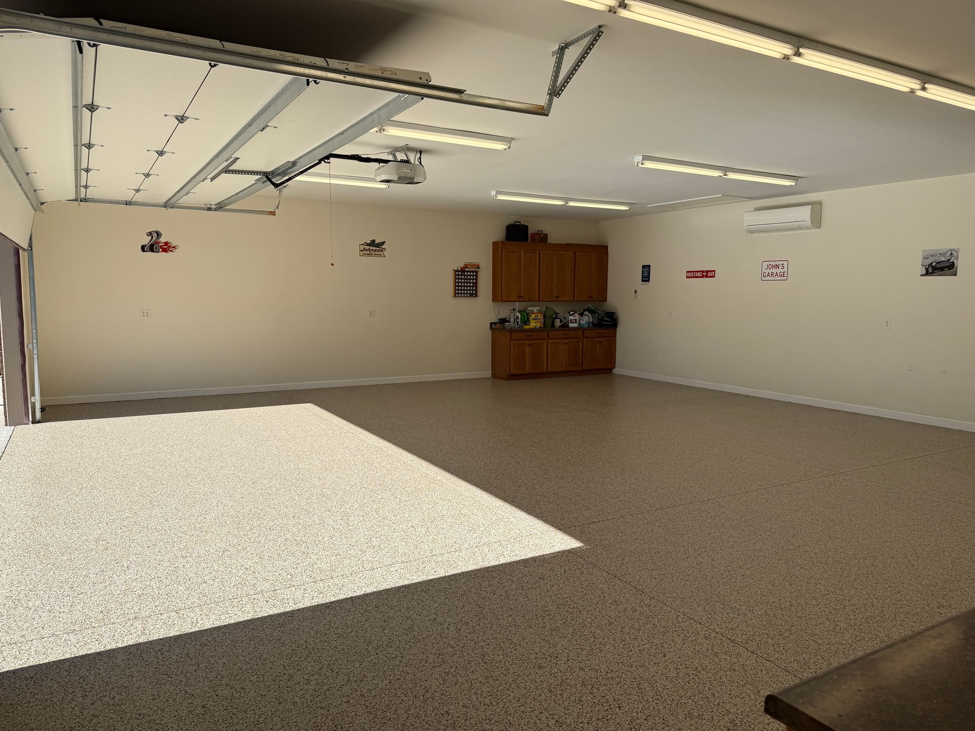 Empty garage with beige walls, speckled floor, fluorescent lights, and a wooden cabinet against the back wall.