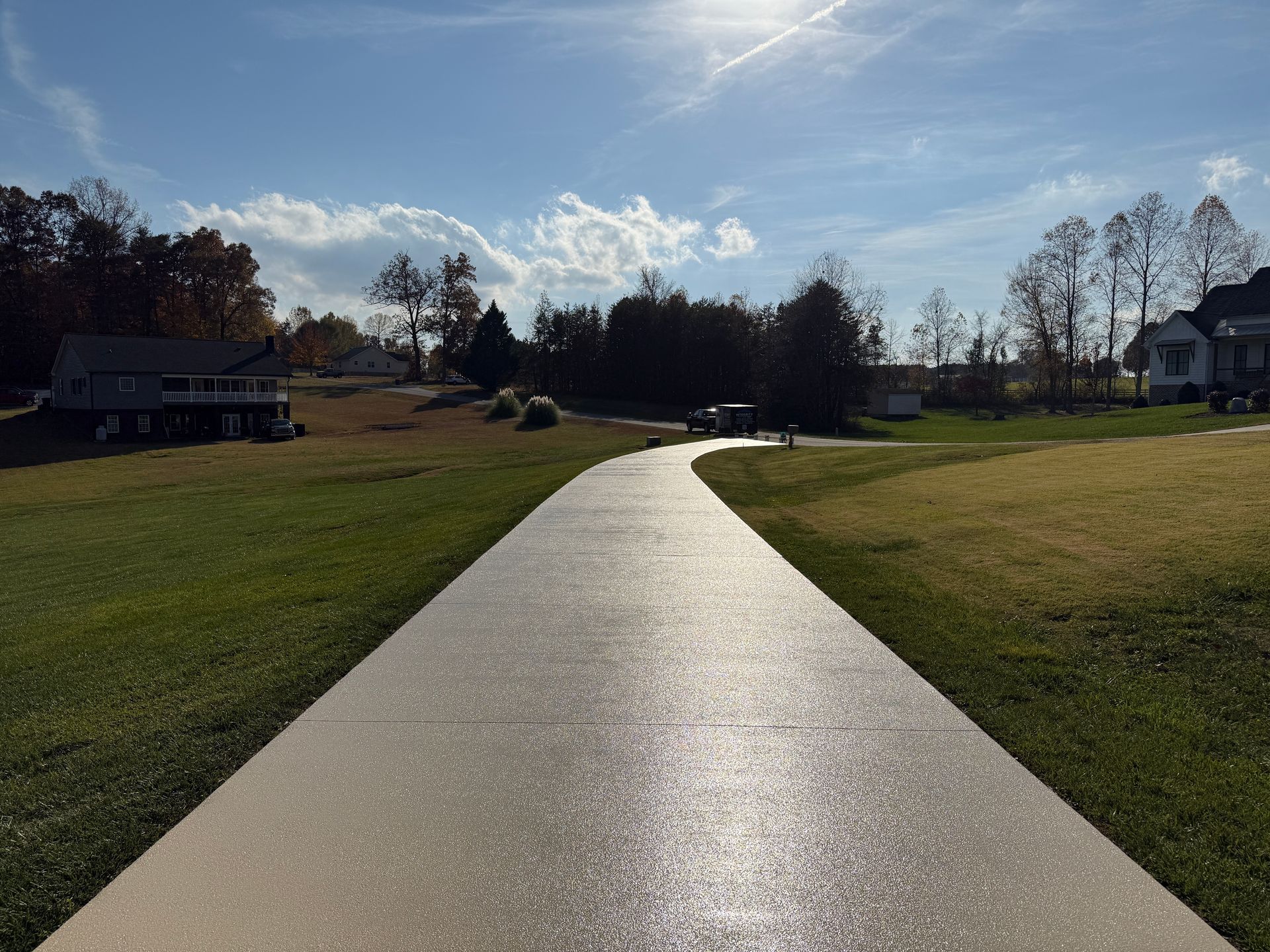 Curved concrete path through a grassy park under a bright blue sky with trees and houses in the distance