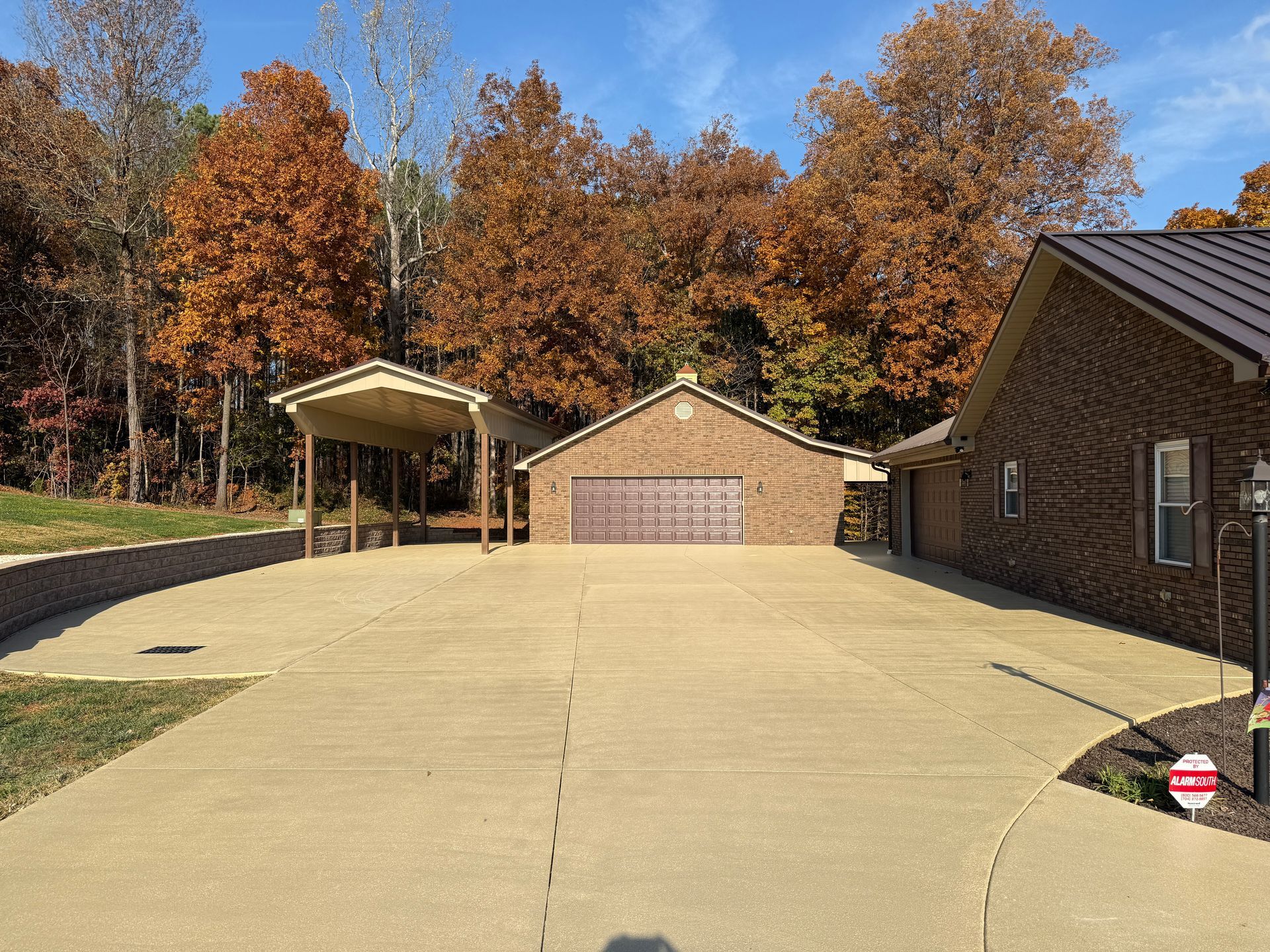 Wide concrete driveway leading to a brick garage beside a house, with autumn trees in the background