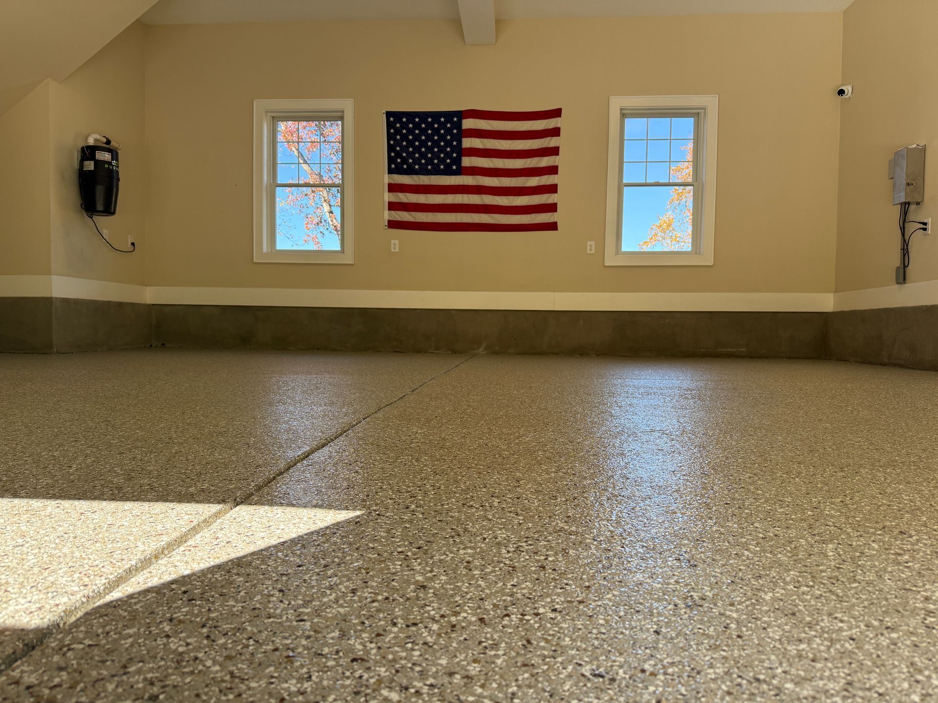 Empty garage with speckled floor, beige walls, two windows, and a U.S. flag on the back wall.