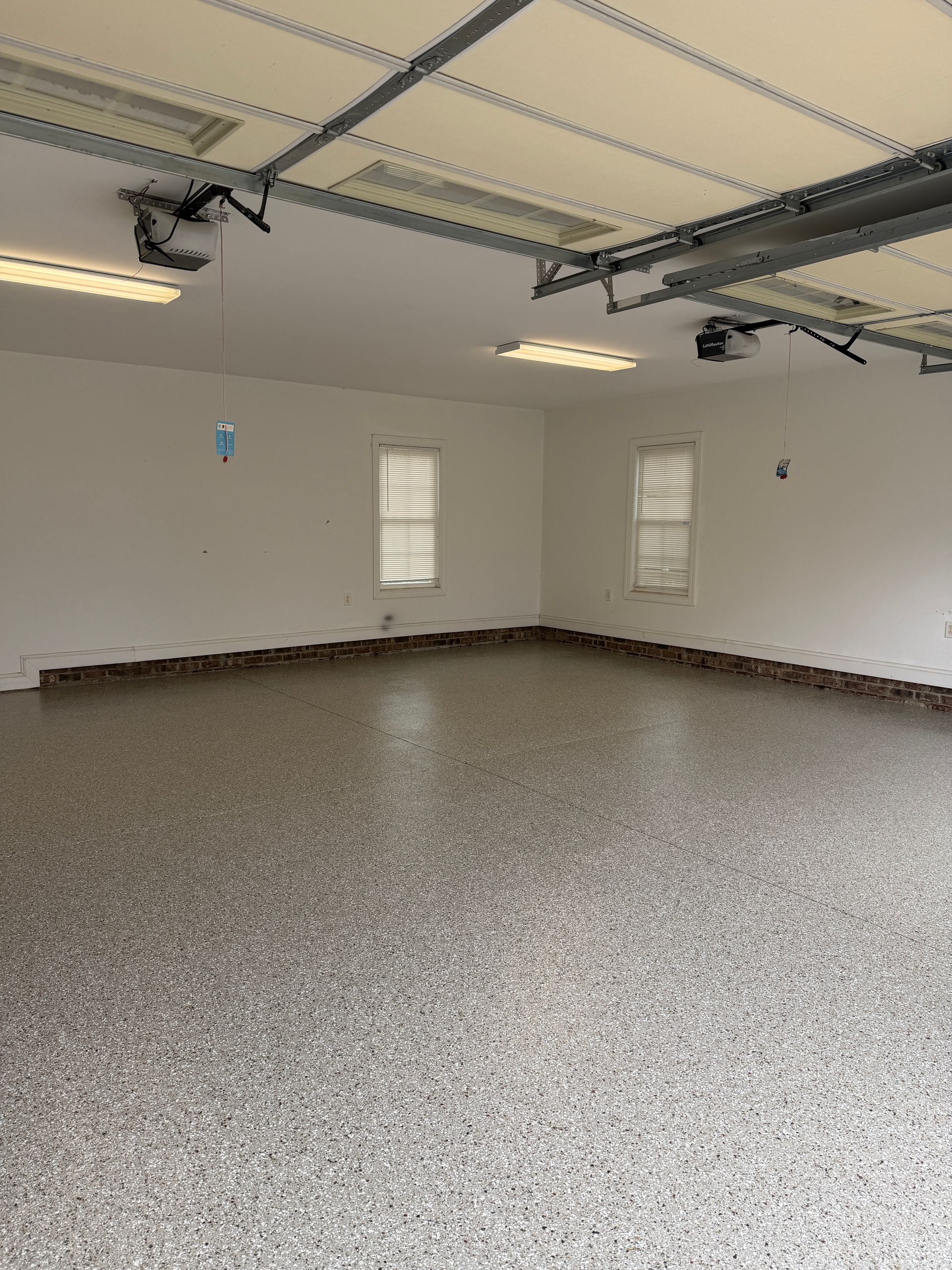 Empty garage with speckled concrete floor, white walls, overhead door tracks, and windows on the back wall