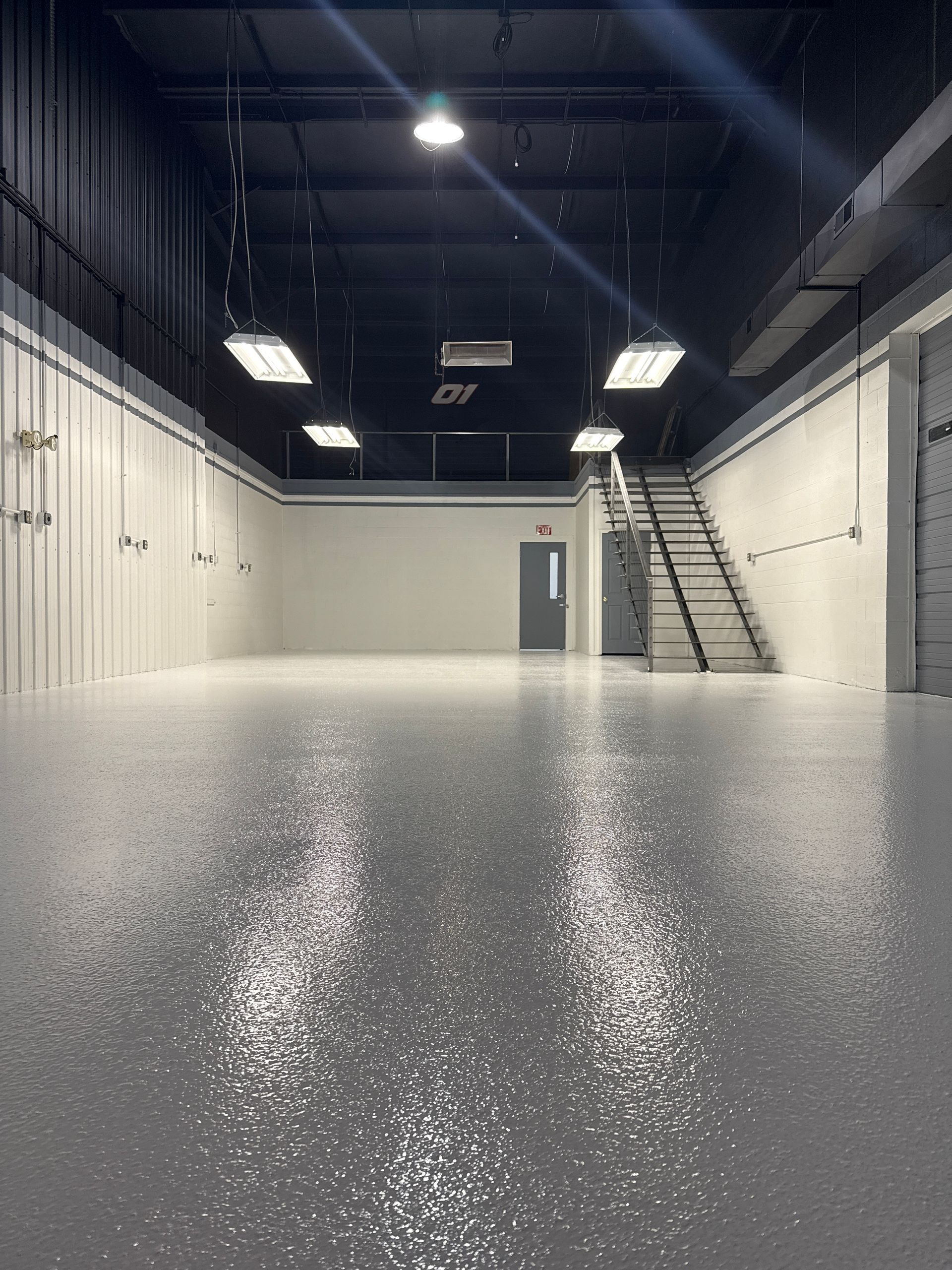Empty industrial room with white walls, polished floor, high ceiling lights, and a metal staircase.
