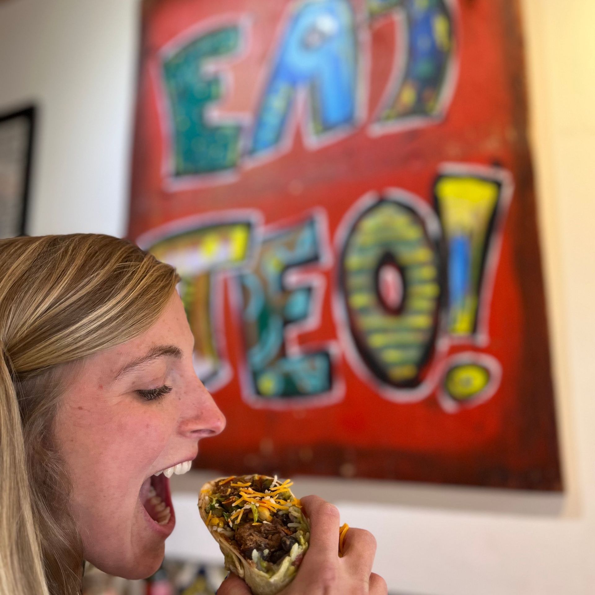 A woman is eating a burrito in front of a sign that says eat teo