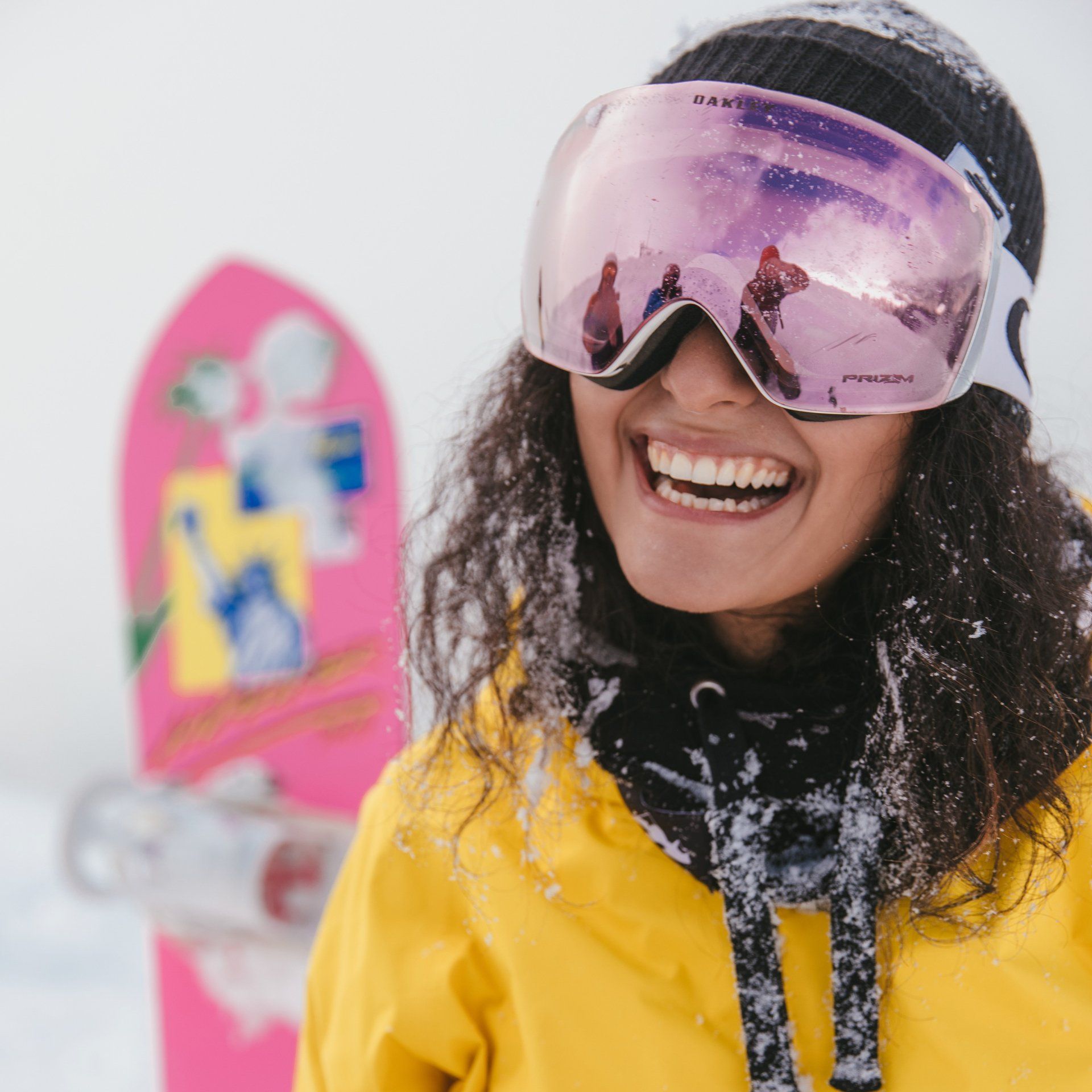 A woman wearing goggles is smiling in front of a pink snowboard