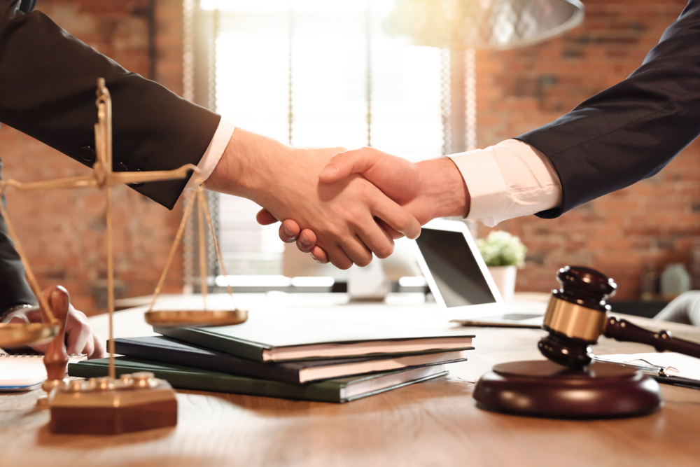 Two professionals shaking hands across a desk with legal documents and a gavel in a courtroom-like office.