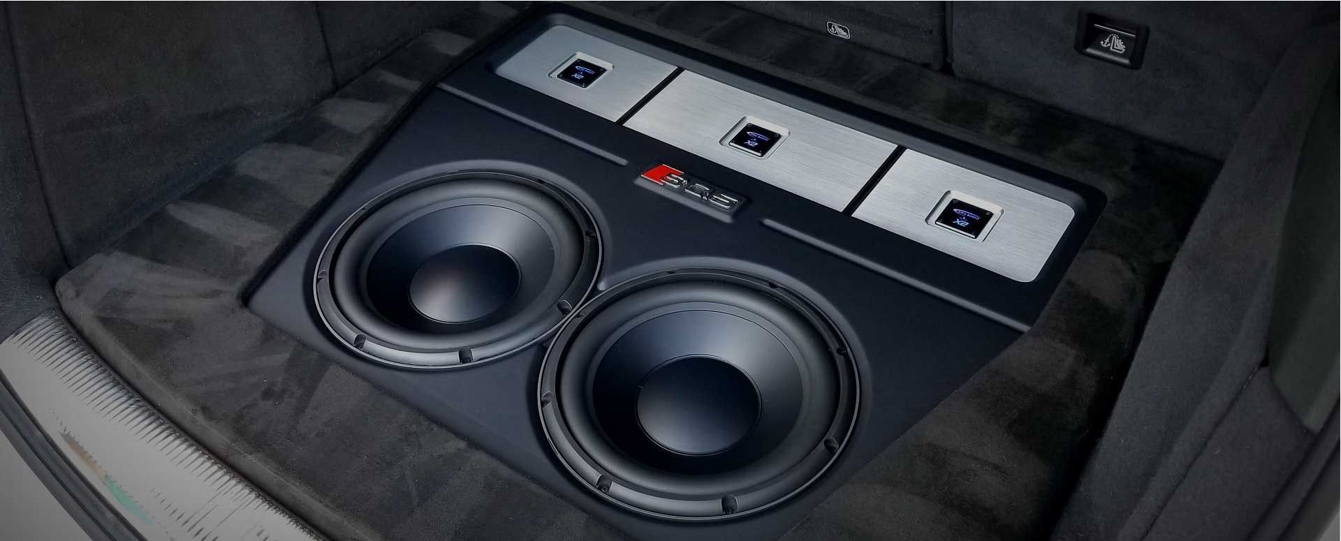 Car's trunk with a custom black subwoofer box, holding two large speakers and amplifier.