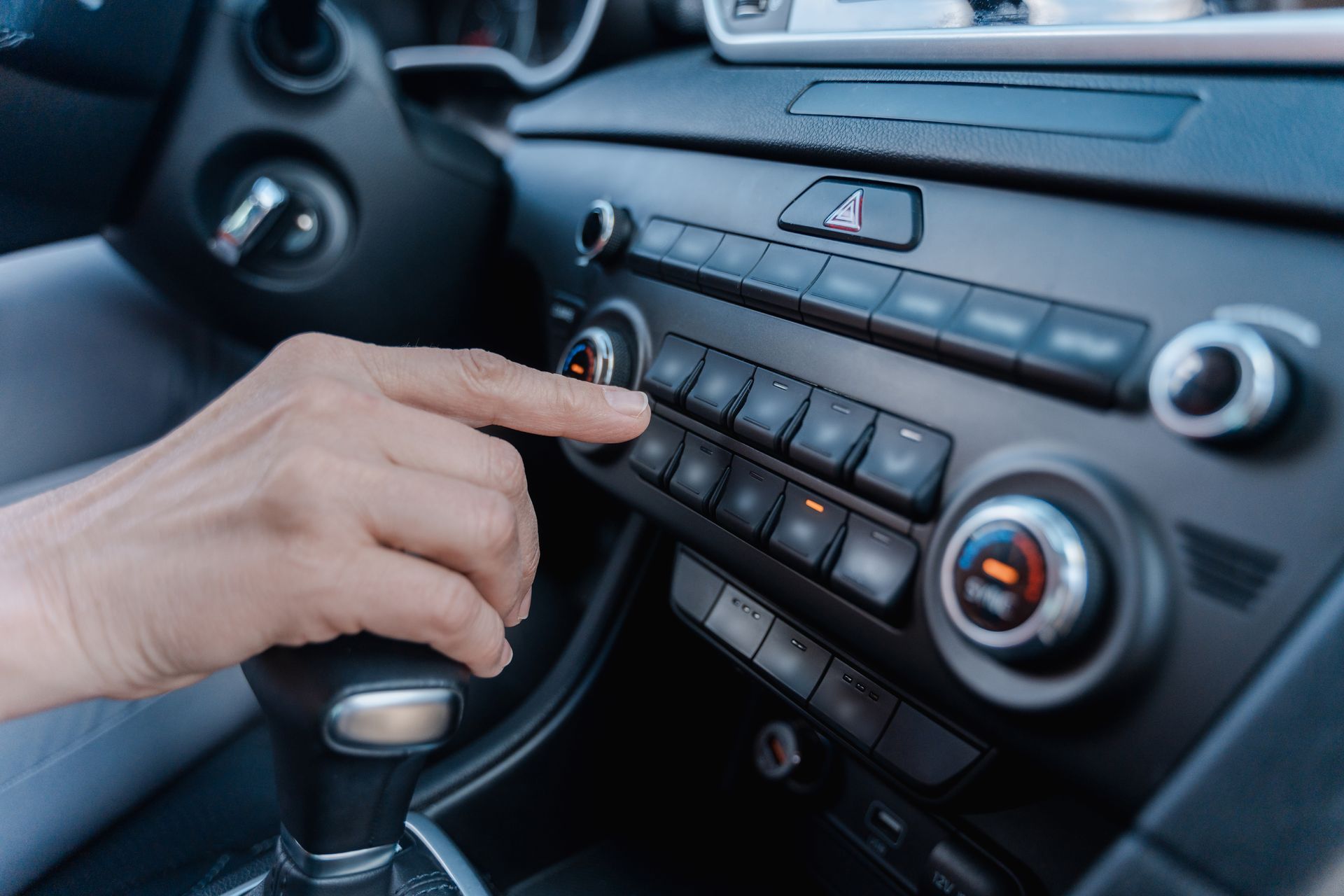 Person's hand pressing a button on a car's dashboard with various controls and gear shift visible.