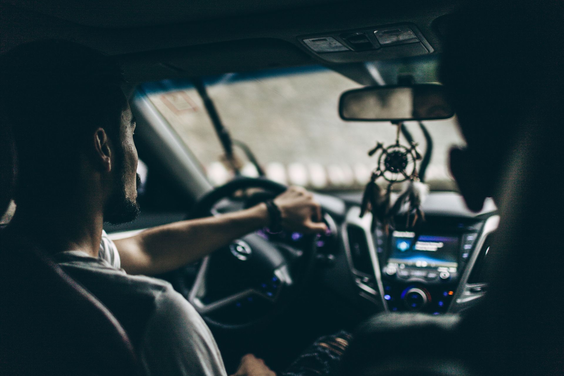 Person driving a car, hands on the steering wheel, seen from the back seat. Dashboard and rear-view mirror are visible.