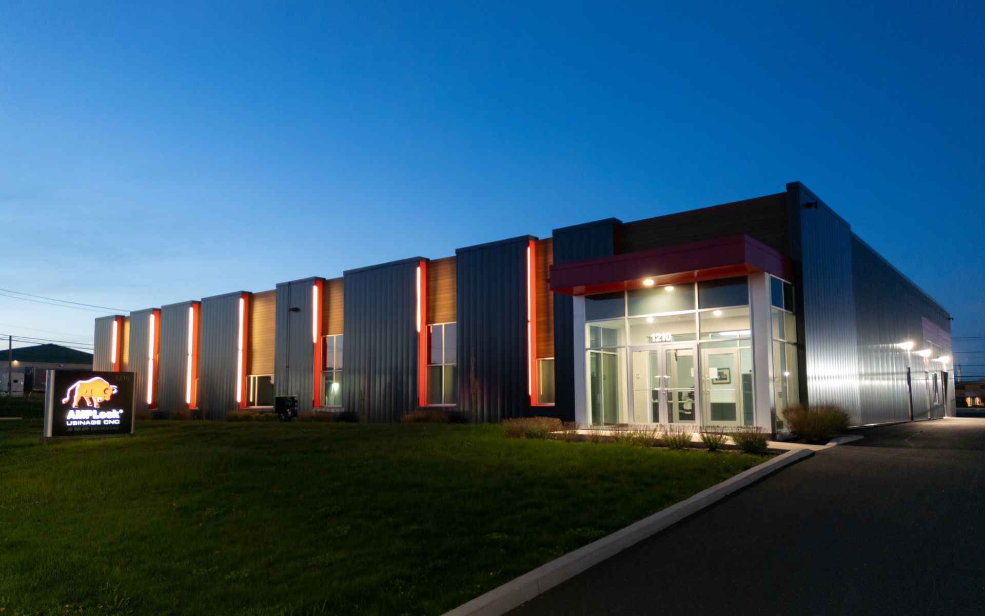 Bâtiment commercial moderne au crépuscule, avec des rayures verticales orange illuminées, un logo et une entrée vitrée.