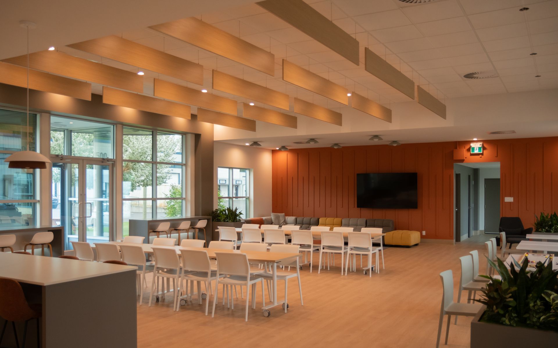 Pièce moderne ouverte avec tables et chaises, un grand écran et des touches d'orange, avec une entrée donnant sur l'extérieur.